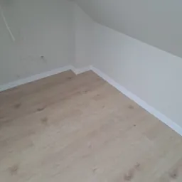 Jasne panele podłogowe ułożone w pokoju na poddaszu, widoczne białe listwy przypodłogowe i fragment instalacji elektrycznej na ścianie.