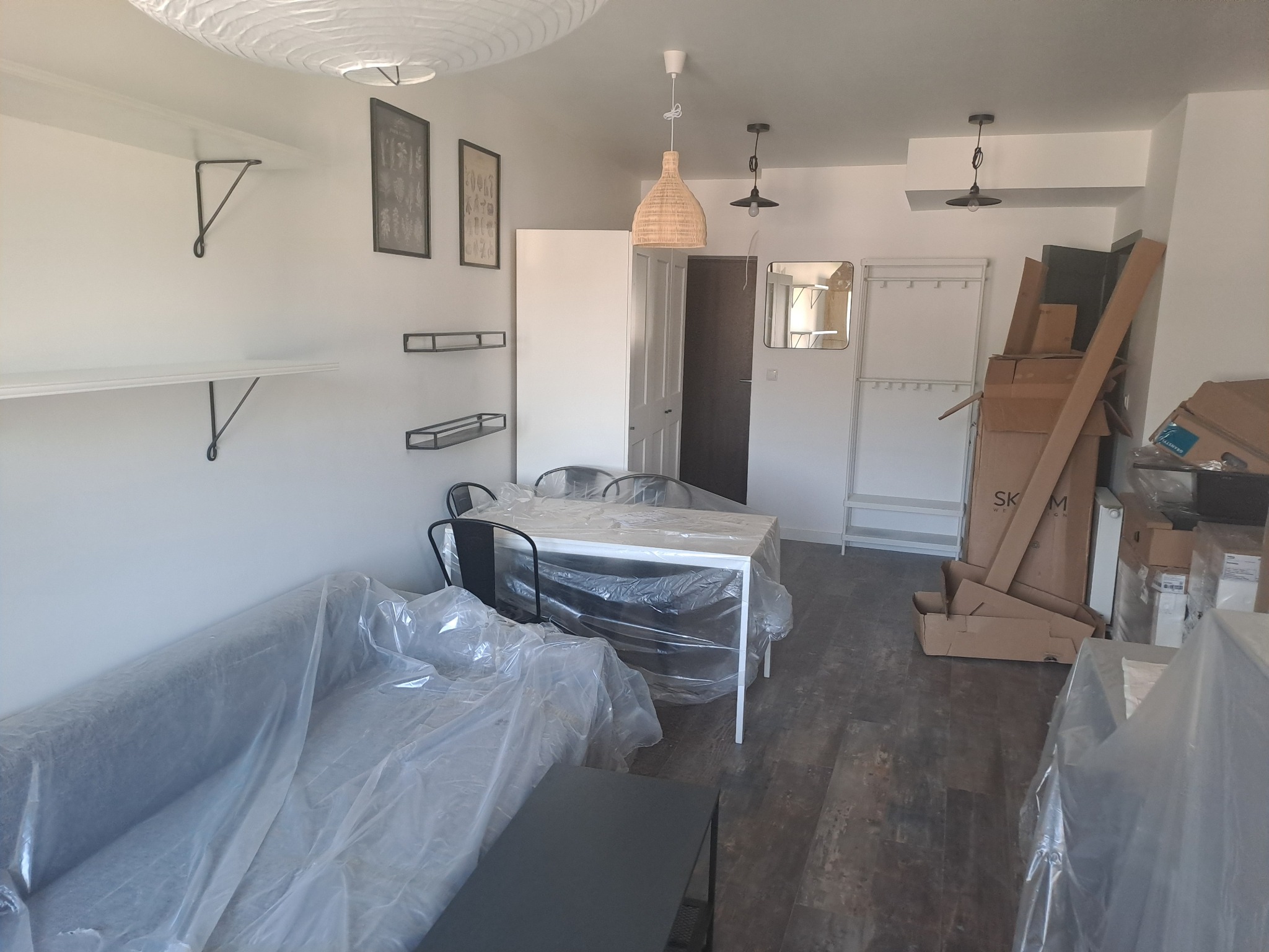 Przygotowania do remontu mieszkania: meble i podłoga zabezpieczone folią ochronną, kartony ze sprzętem i materiałami budowlanymi w tle, widoczne półki ścienne i lampy sufitowe.