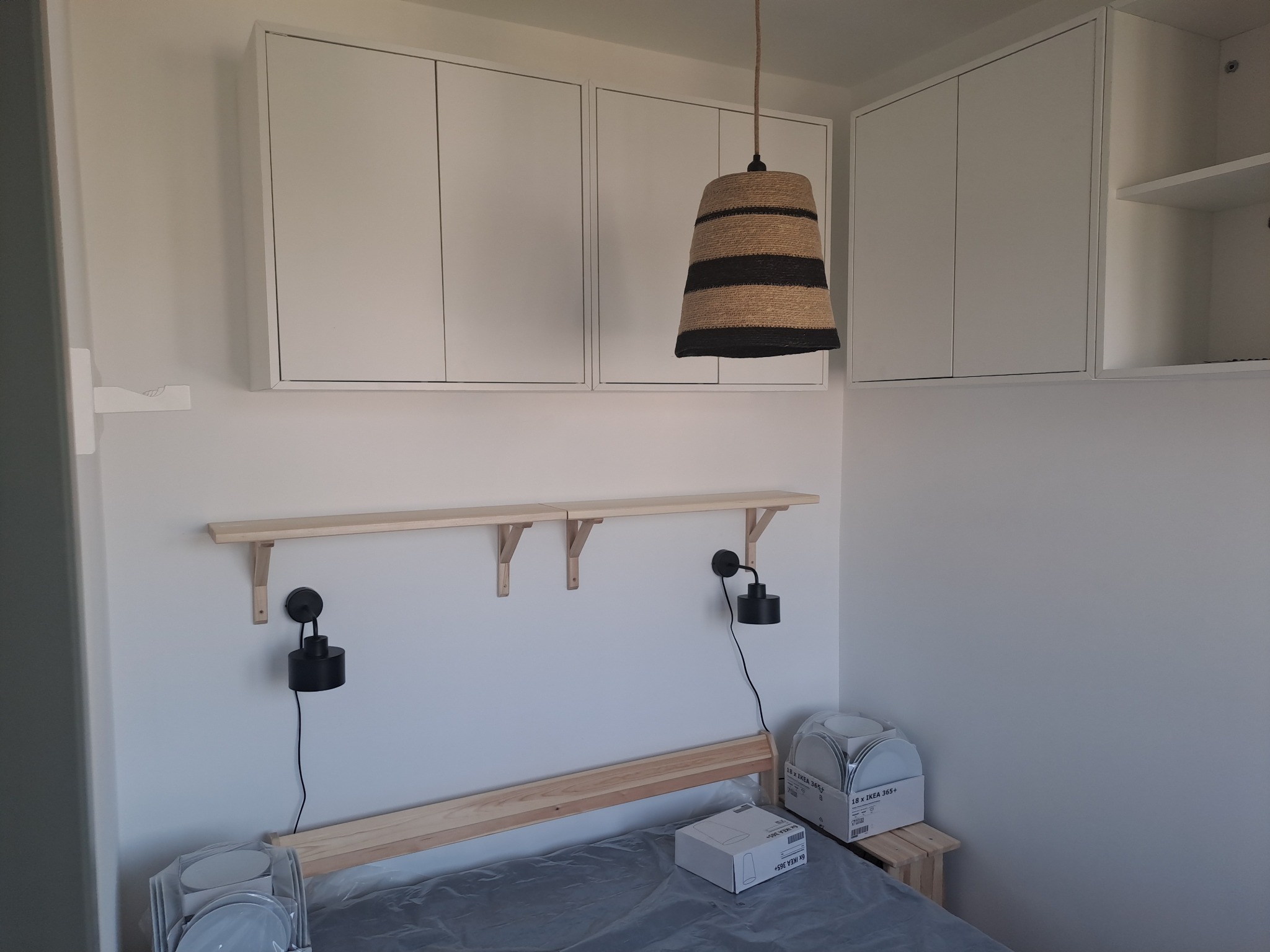 Nowoczesna sypialnia z drewnianym łóżkiem, półkami i szafkami ściennymi w białym kolorze, oświetlona lampą wiszącą z abażurem z naturalnego sznurka.