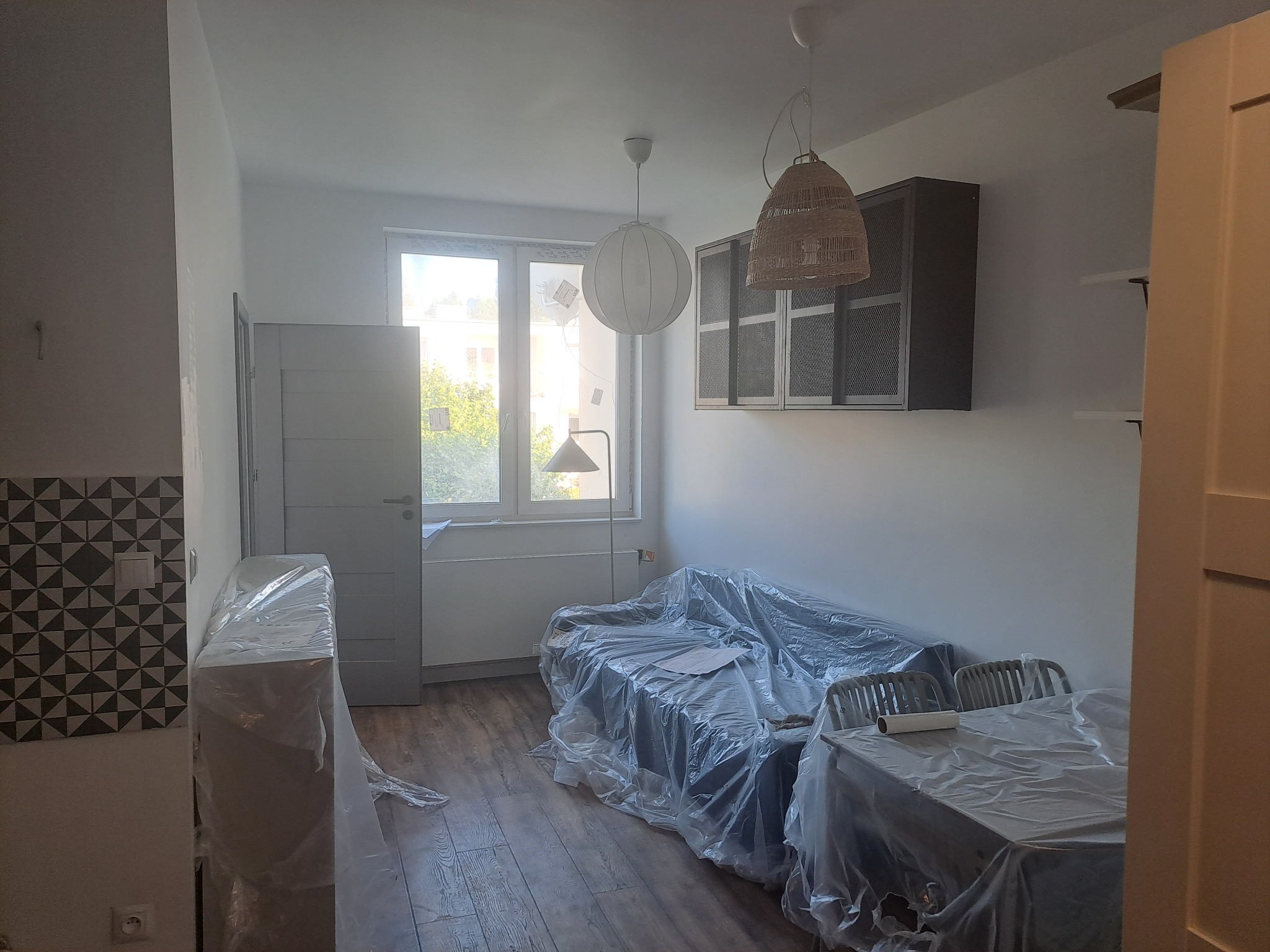Pokój w trakcie przygotowań do malowania: meble i podłoga zabezpieczone folią malarską, widoczne lampy wiszące i wzorzysta ściana w tle.
