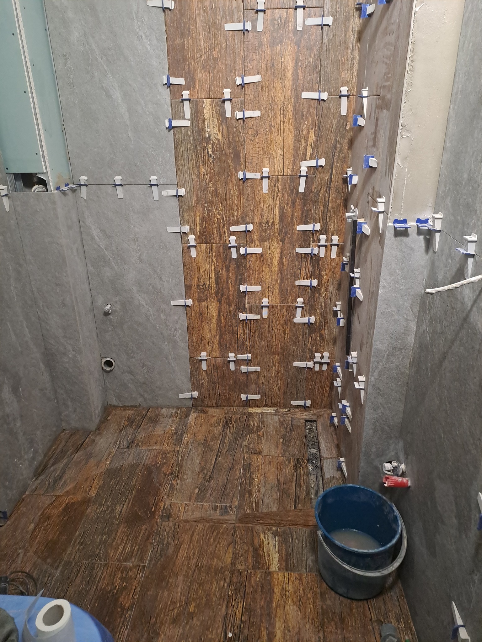 Układanie płytek imitujących drewno na podłodze i ścianie łazienki, widoczne plastikowe krzyżyki dystansowe, wiadro z wodą i gąbka. Szare płytki na sąsiedniej ścianie.