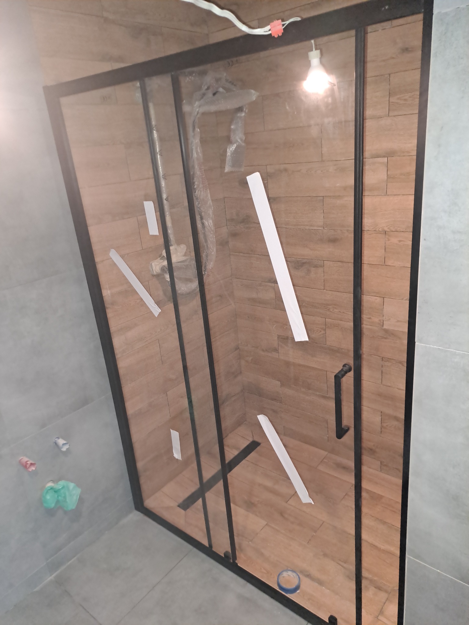 Kabina prysznicowa z czarnymi profilami, zamontowana w łazience z szarymi i drewnopodobnymi płytkami, zabezpieczona taśmą malarską w trakcie prac wykończeniowych.