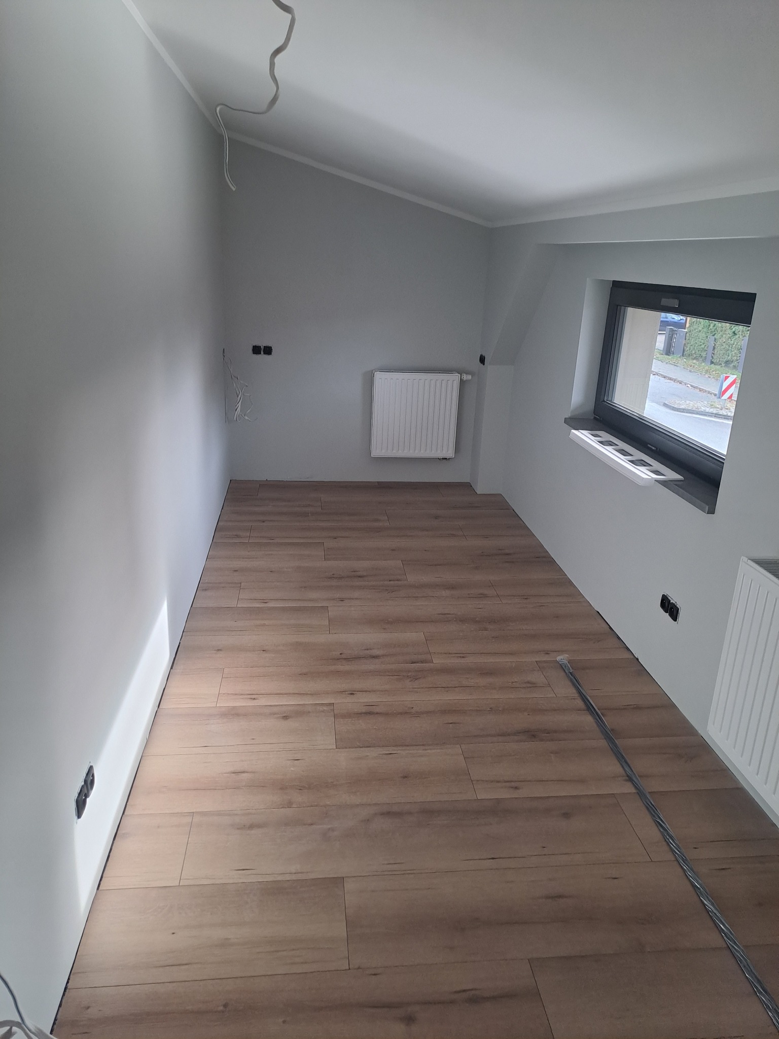 Pokój po remoncie z ułożonymi panelami podłogowymi w odcieniach brązu, szare ściany z widocznymi gniazdkami elektrycznymi, grzejnik i okno z widokiem na zieleń za oknem. Na podłodze leży listwa...