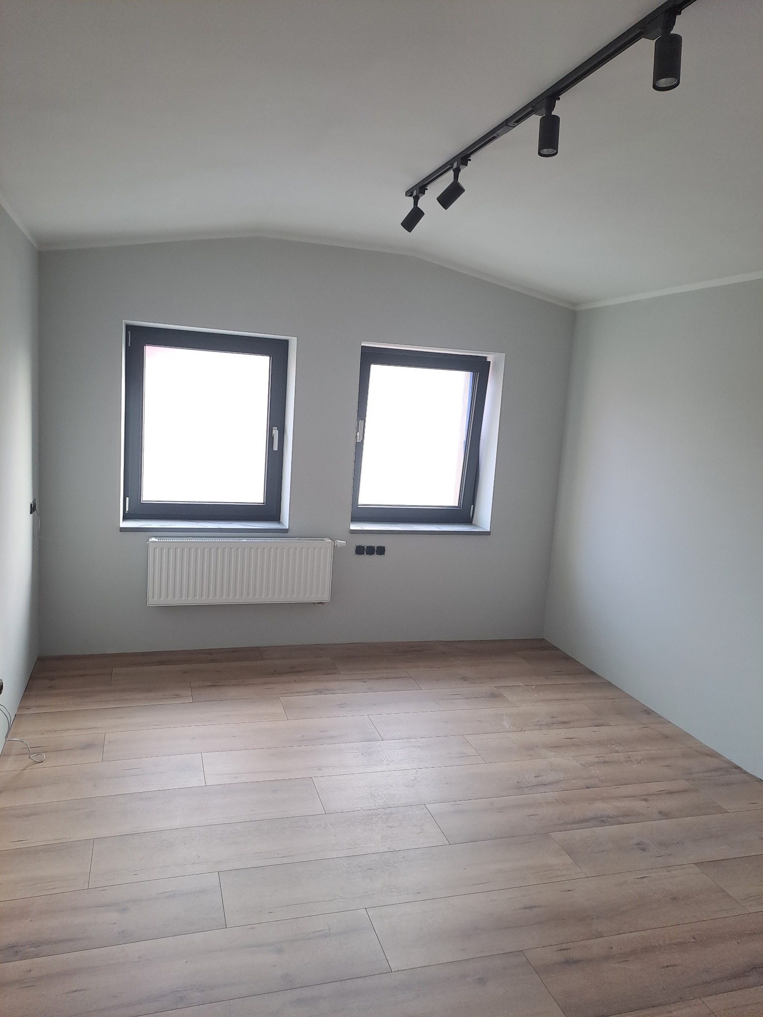 Pusty pokój po remoncie z panelami podłogowymi w odcieniach brązu, szare ściany, dwa okna z czarnymi ramami, biały grzejnik pod oknem i czarna listwa z czterema reflektorami na suficie.