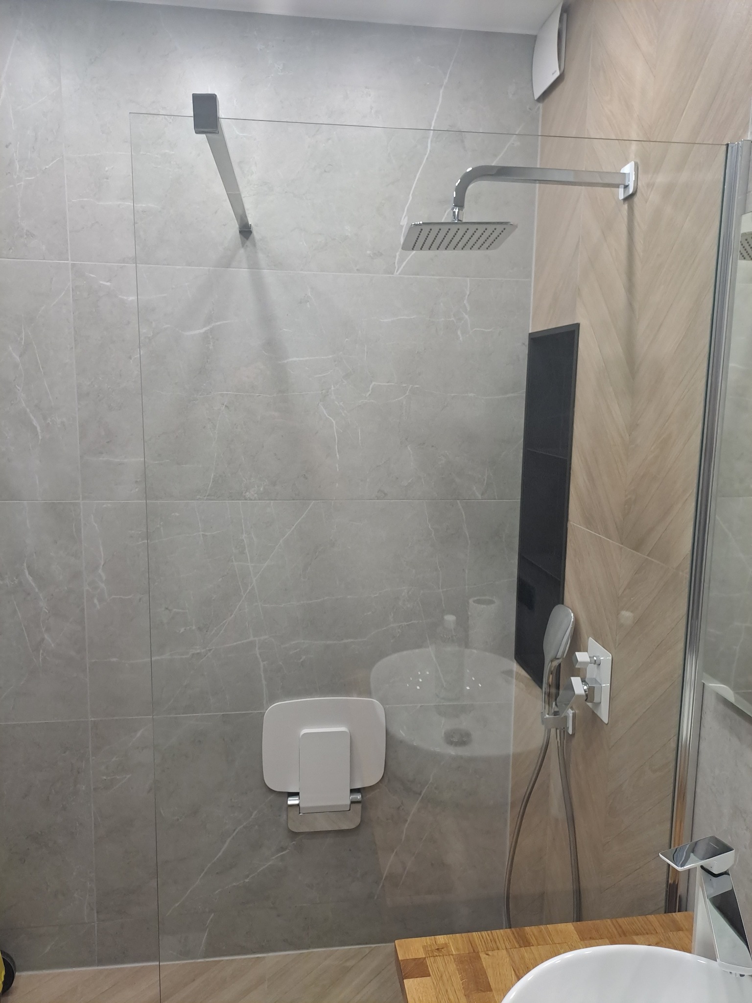 Nowoczesna kabina prysznicowa z deszczownicą i siedziskiem składanym, wykończona płytkami imitującymi marmur, z widoczną umywalką i baterią.