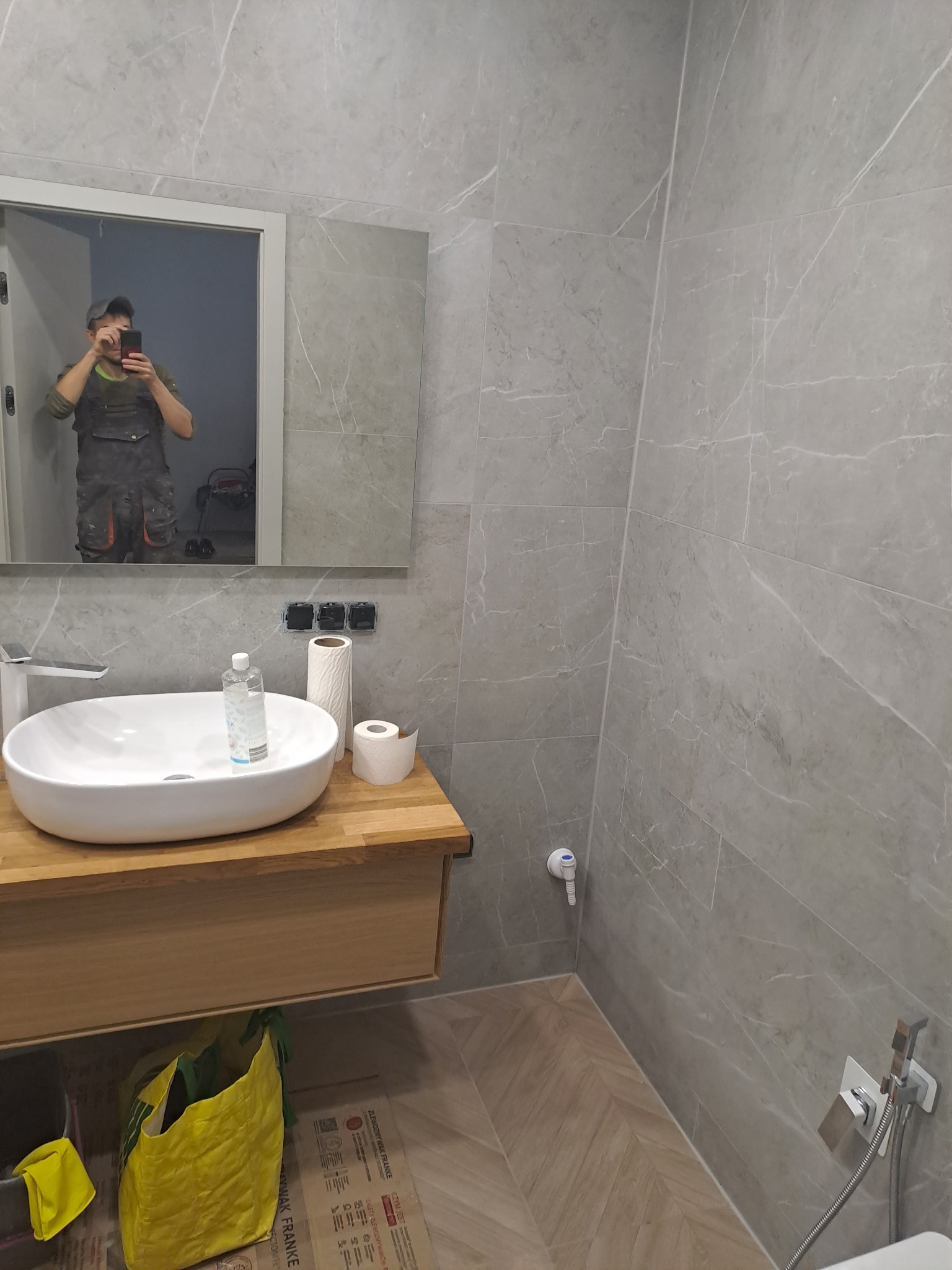 Nowoczesna łazienka z szarymi płytkami imitującymi marmur, drewnianą szafką pod umywalką i widocznym odbiciem osoby robiącej zdjęcie w lustrze.