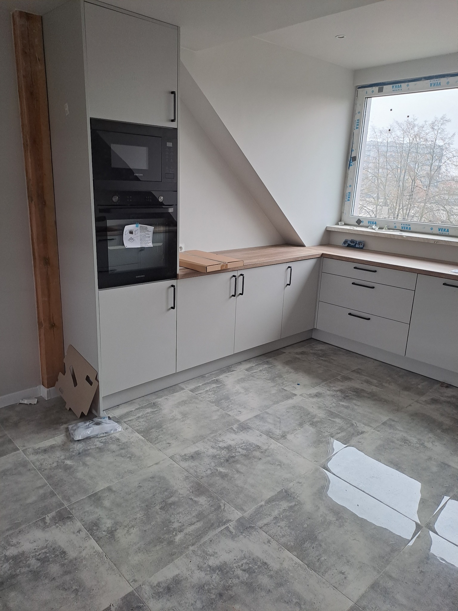Nowoczesna kuchnia w trakcie wykańczania pod skosem dachu, z szarymi szafkami, drewnianym blatem i szarą płytką podłogową imitującą kamień. Widoczne kartony po sprzęcie AGD i ślady po pracach...