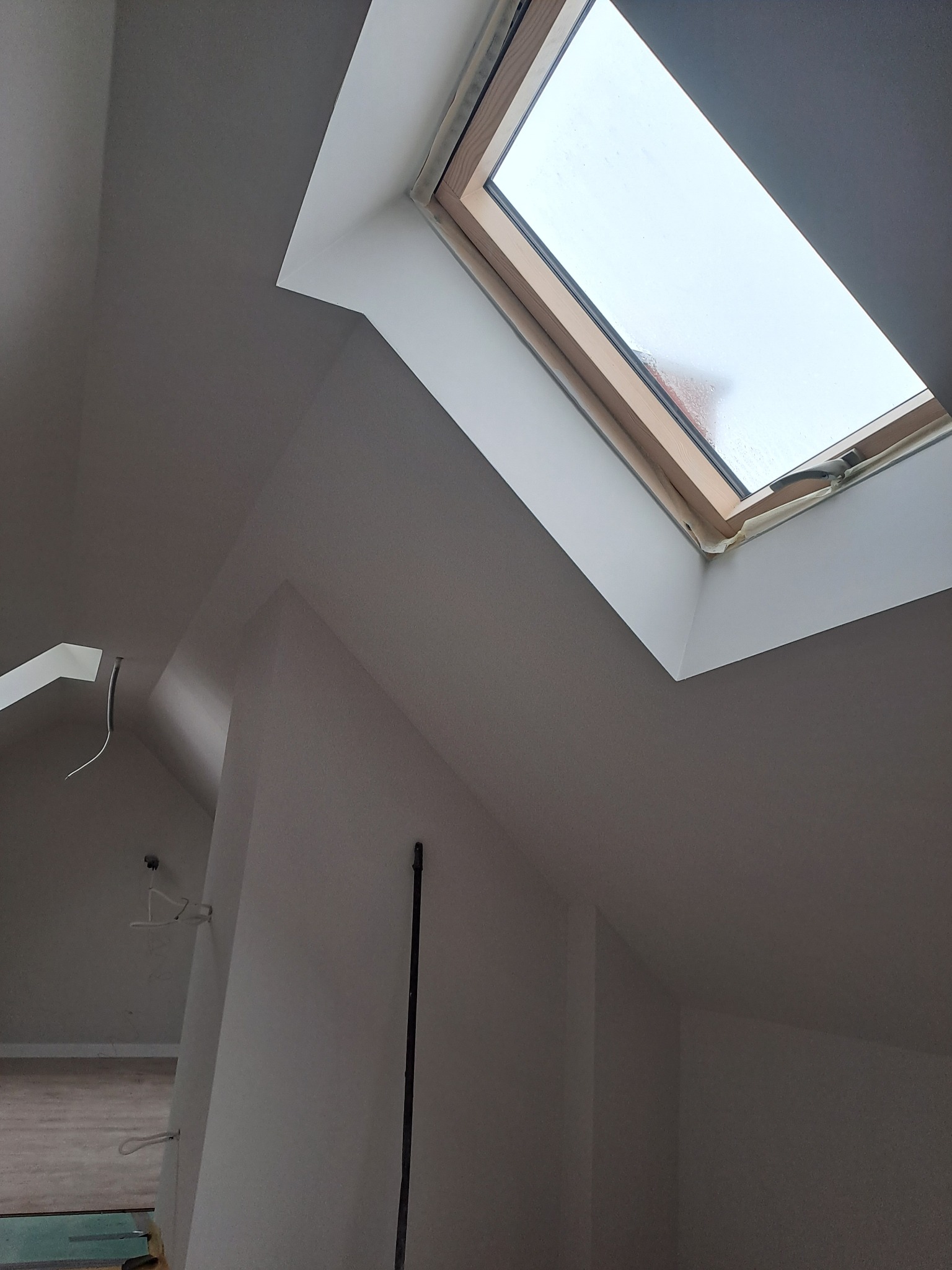 Poddasze po malowaniu na biało, widoczne okno dachowe z drewnianą ramą, przewody elektryczne wystające ze ściany, fragment podłogi z zabezpieczeniem.