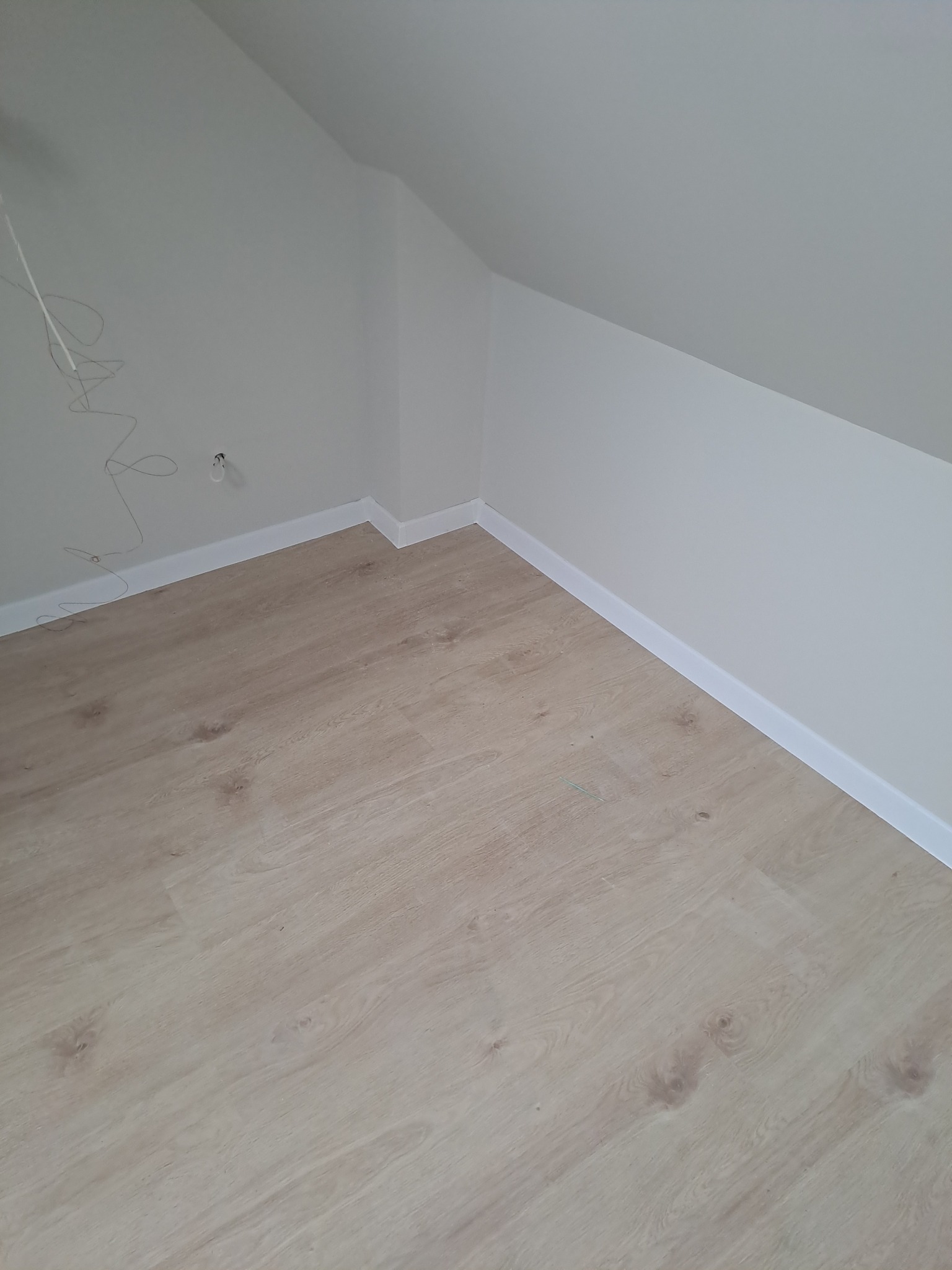 Jasne panele podłogowe ułożone w pokoju na poddaszu, widoczne białe listwy przypodłogowe i fragment instalacji elektrycznej na ścianie.