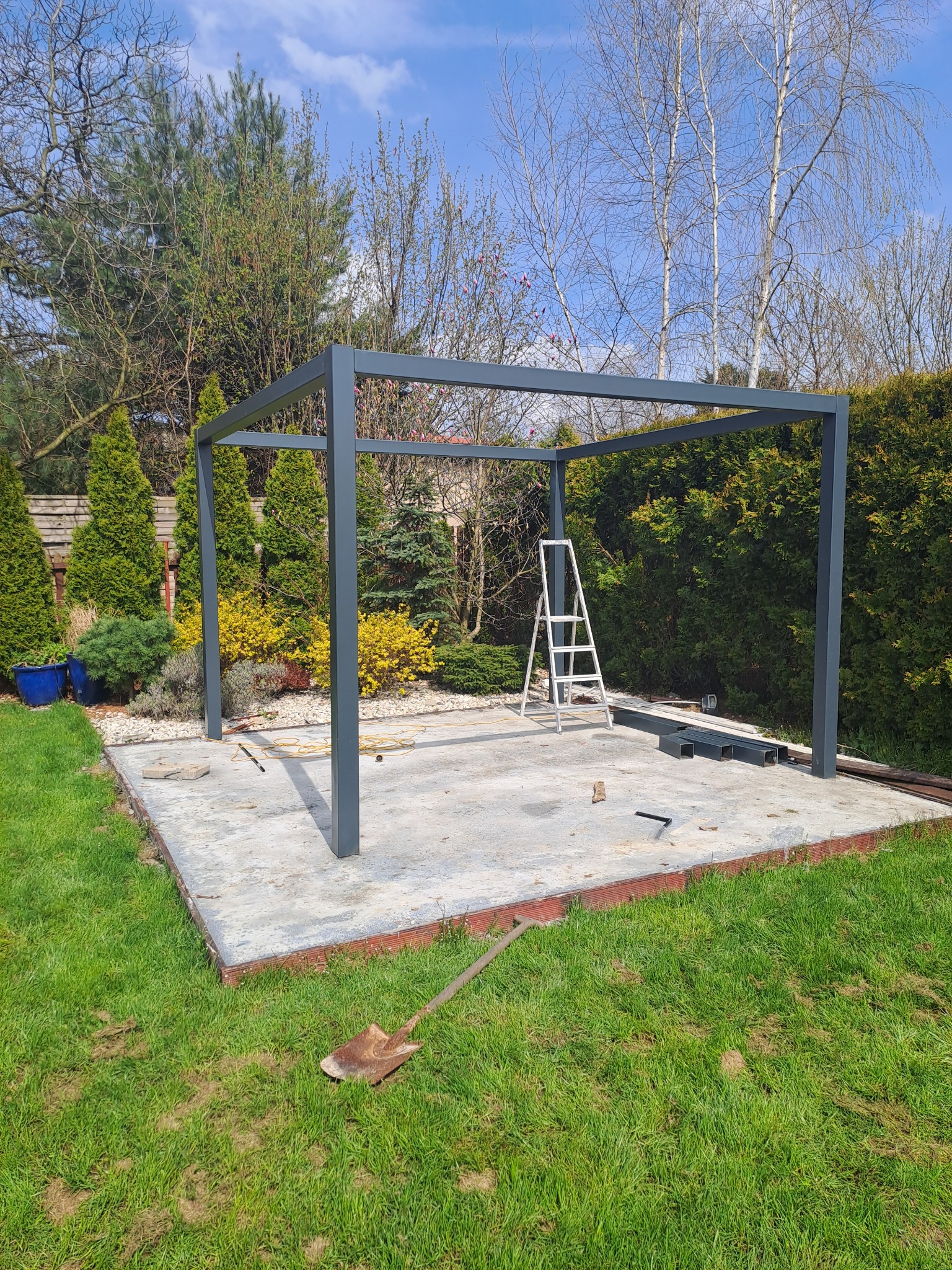 Konstrukcja altany ogrodowej w trakcie montażu, widoczna drabina i narzędzia, otoczenie: trawnik, krzewy i drzewa.