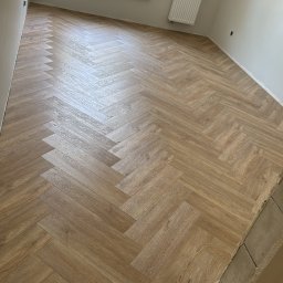 Usługi ogólnobudowlane Bartosz Żaczek - Ułożona podłoga z paneli w jodełkę, widok na okno z parapetem i grzejnik. Na parapecie stoją kubki. Na oknie wisi kurtka. Świeżo wykończone wnętrze.