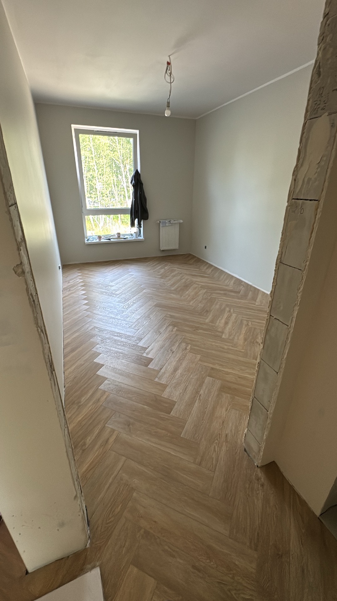 Ułożona podłoga z paneli w jodełkę w jasnym pokoju z oknem. Widoczne fragmenty ścian i niedokończone obramowanie drzwi.