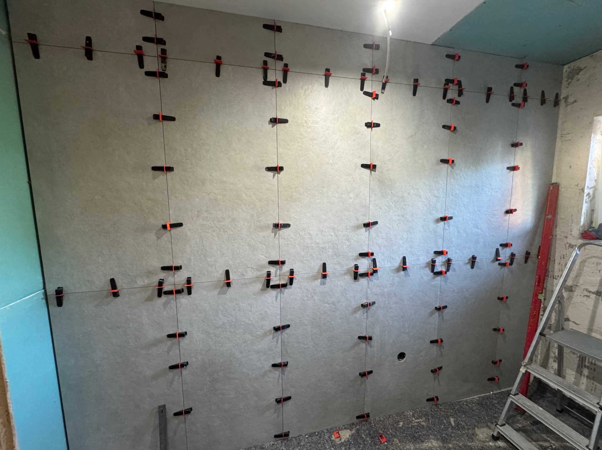 Ściana z szarych płytek, wyrównana systemem poziomowania płytek z czarnymi klipsami i pomarańczowymi klinami, widoczna drabina i poziomica.