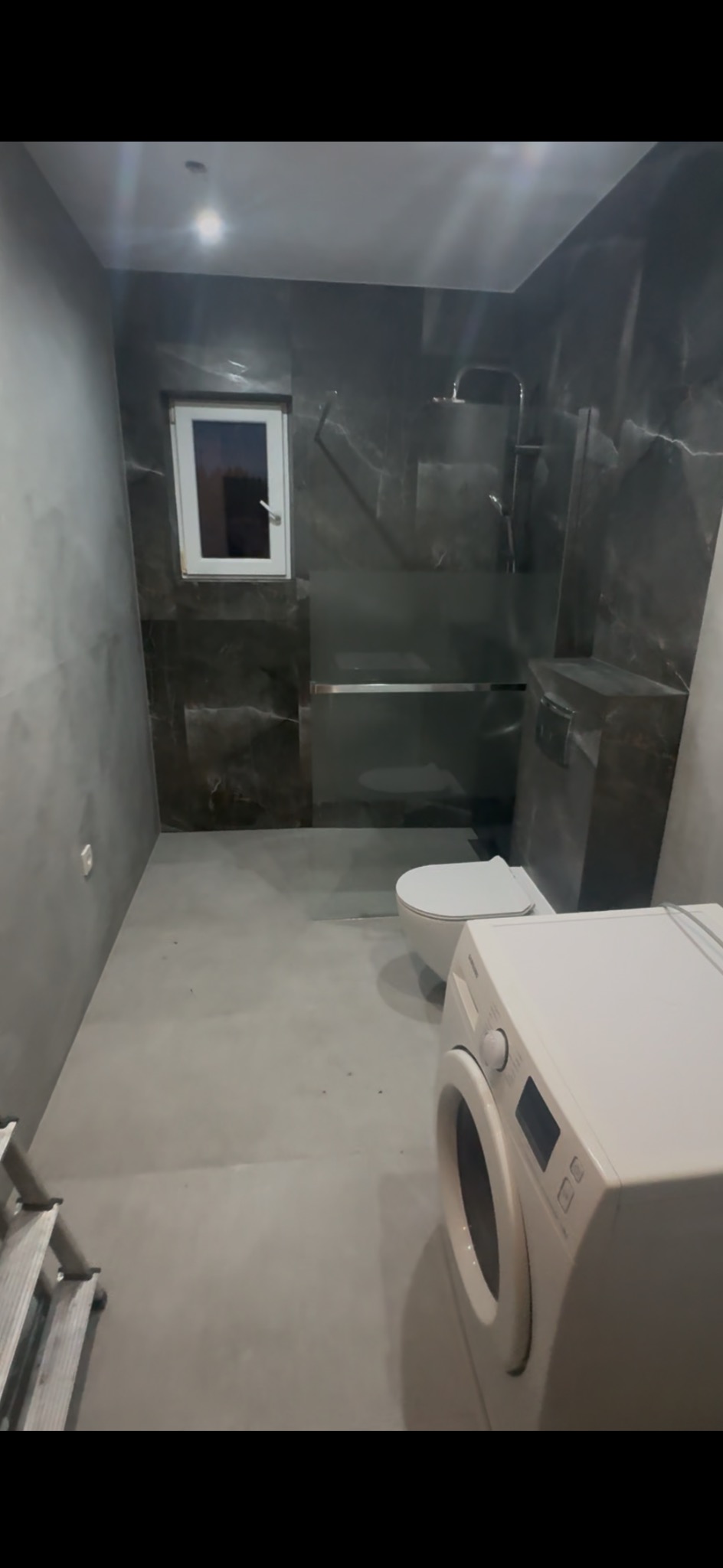 Wykończona łazienka w szarościach z kabiną prysznicową typu walk-in, toaletą i pralką. Widoczne płytki imitujące marmur i gładka posadzka.