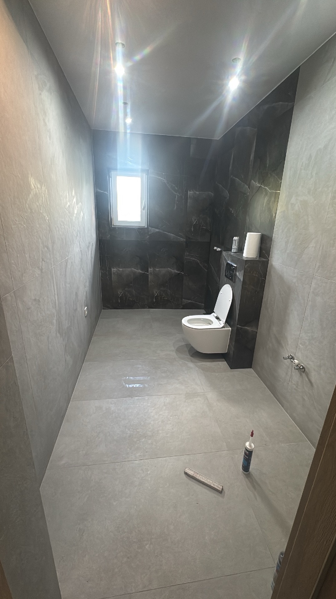 Wykończona łazienka w szarościach: marmurowe płytki na ścianach, szara podłoga, wisząca toaleta. Widoczne akcesoria i narzędzia glazurnicze.