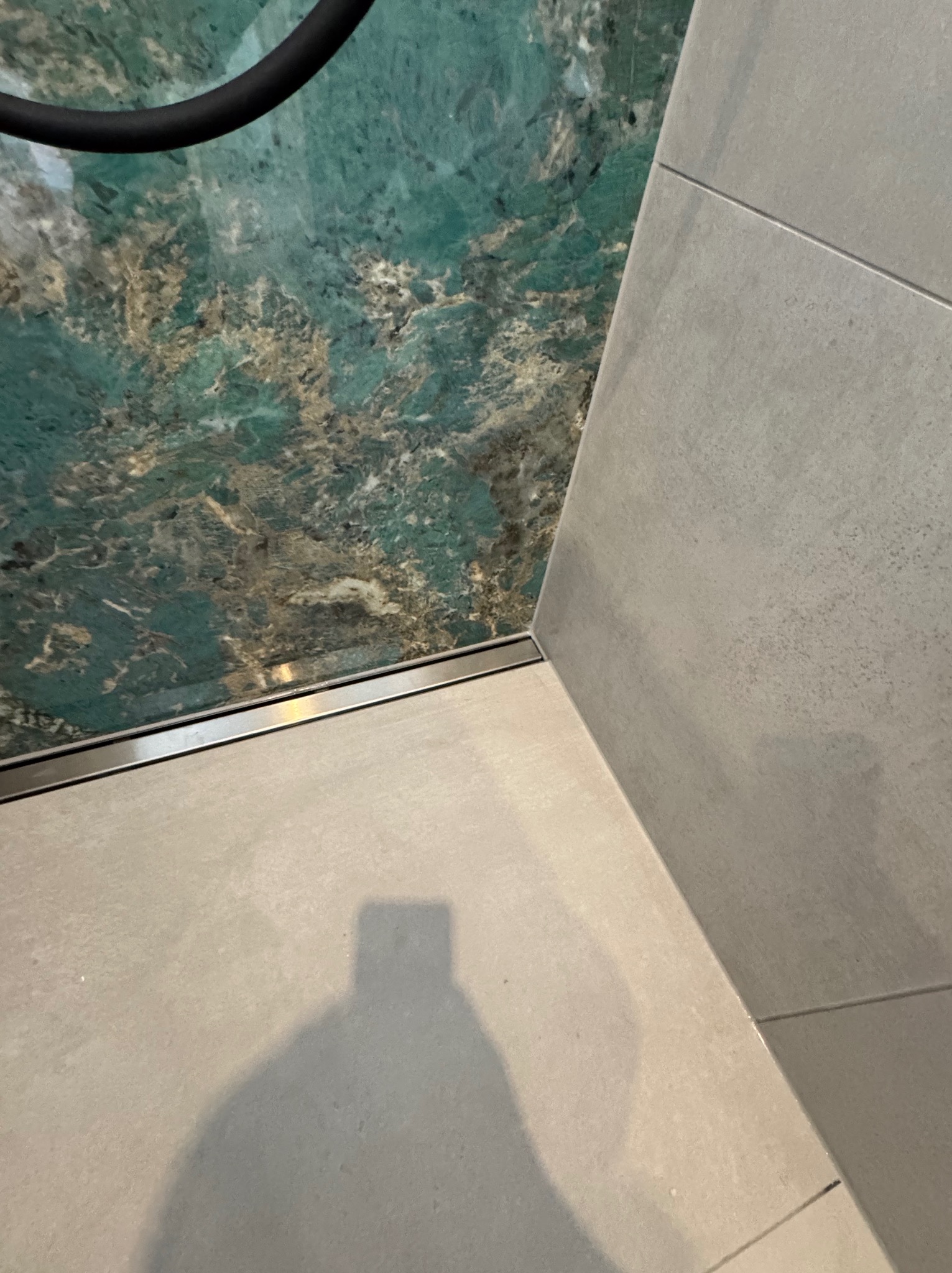 Wykończona strefa prysznicowa z odpływem liniowym, wyłożona płytkami imitującymi marmur w odcieniach zieleni i szarości. Widoczny cień osoby robiącej zdjęcie.