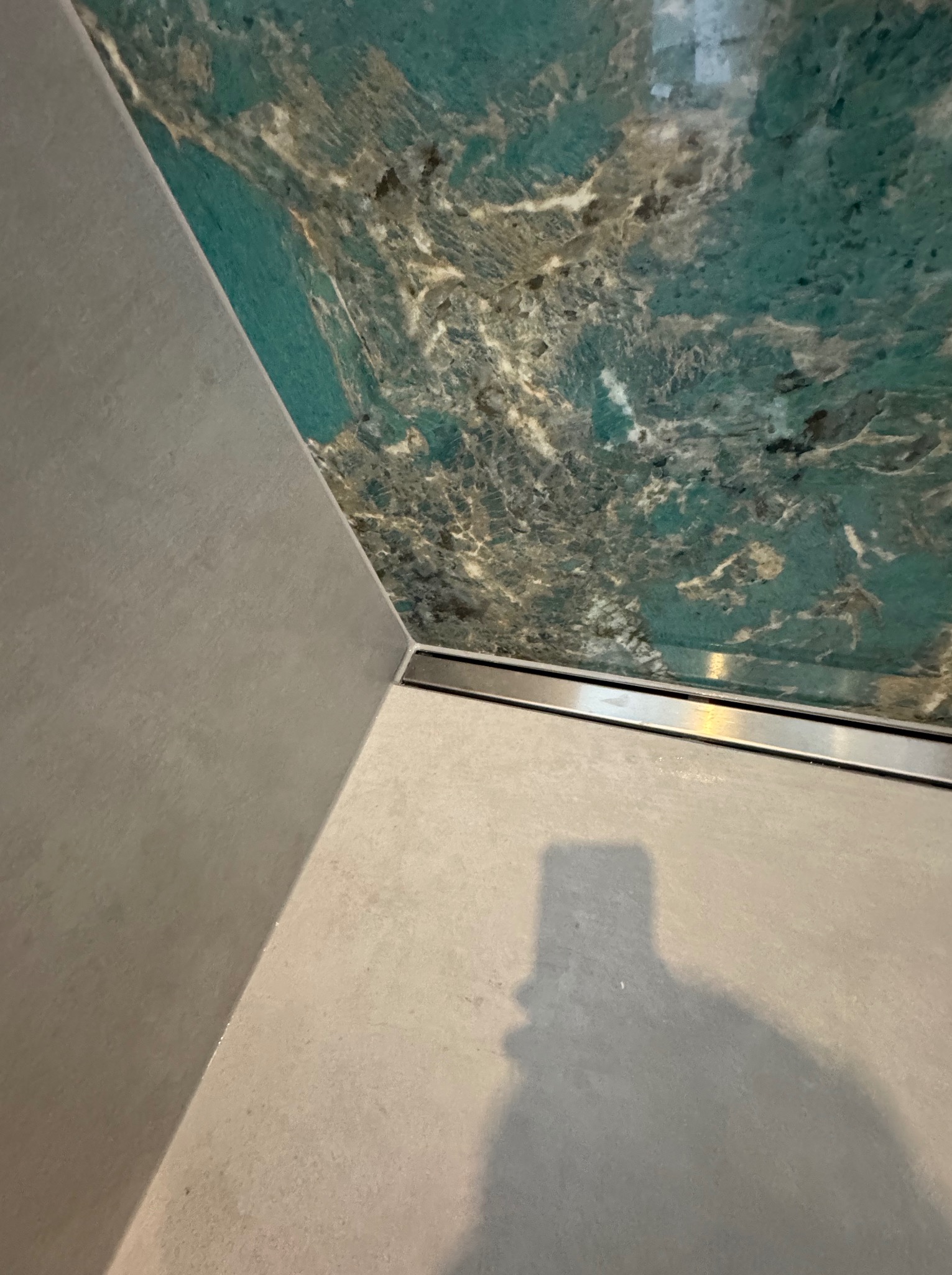 Wykończona łazienka z odpływem liniowym, płytki imitujące marmur na ścianie i podłodze. Minimalistyczny design, widoczny cień osoby robiącej zdjęcie.