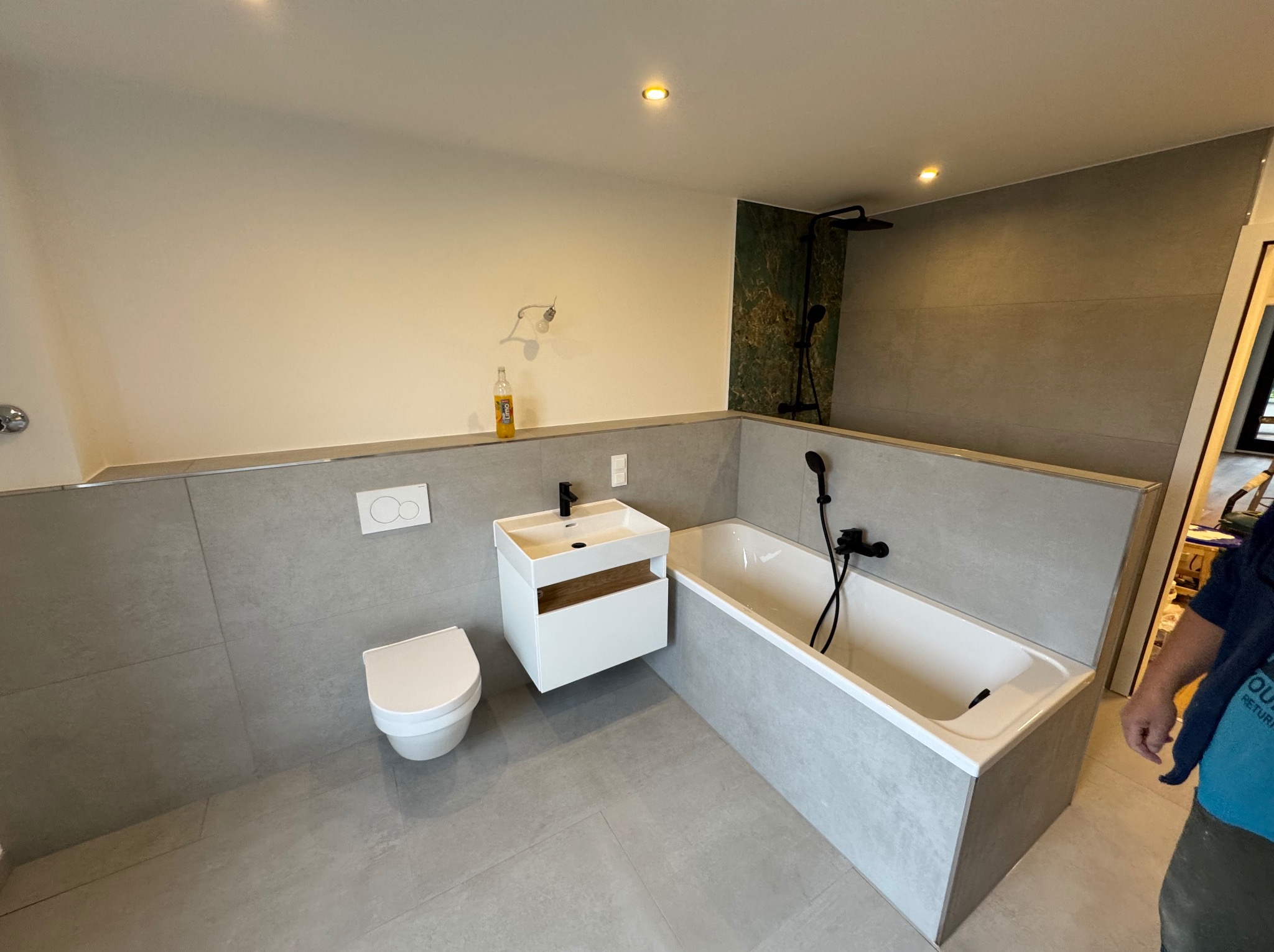 Nowoczesna łazienka z szarą glazurą, białą armaturą i czarnymi detalami. Uwagę zwraca zielona ściana prysznicowa i butelka na ścianie.