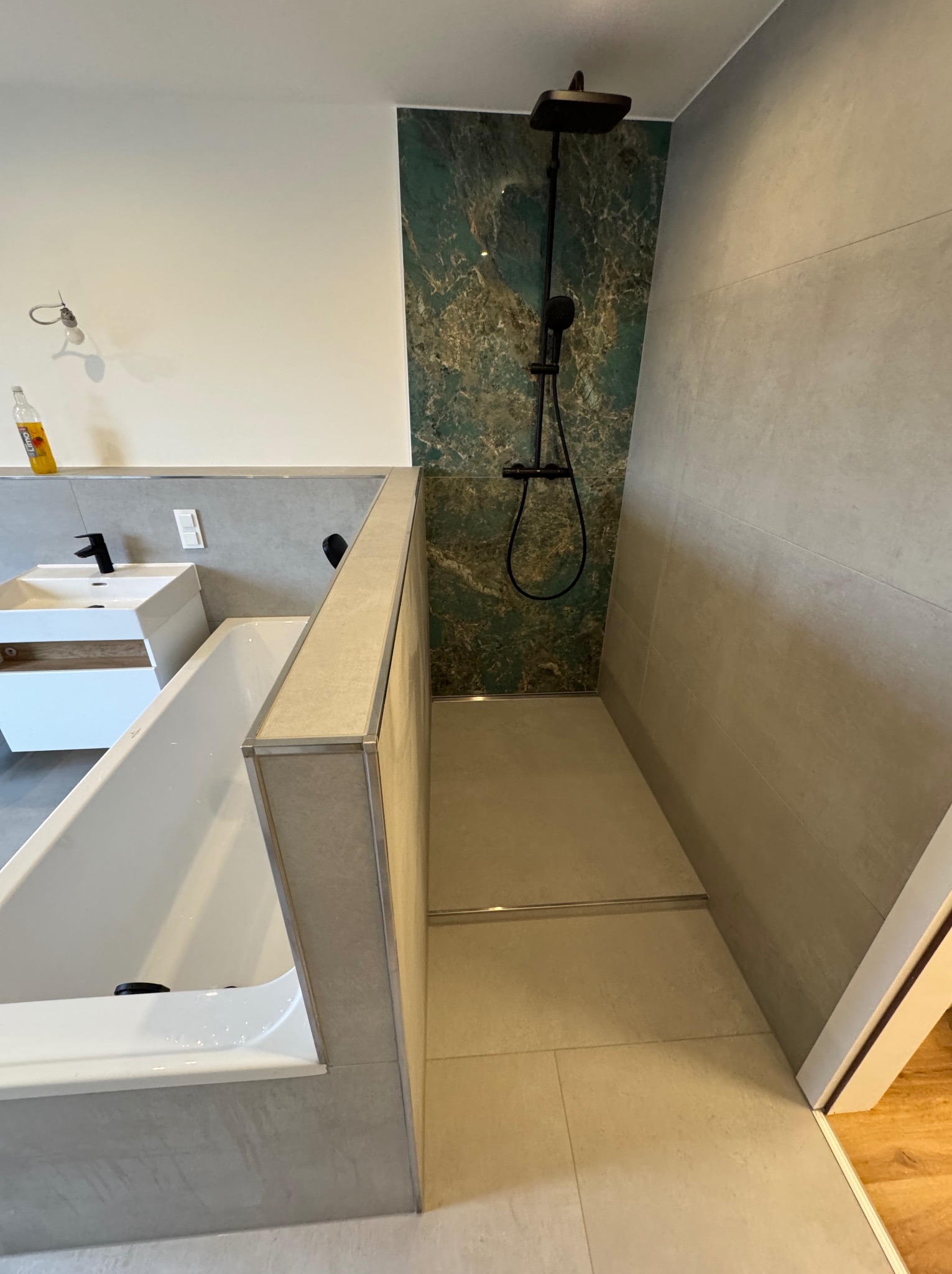 Nowoczesna łazienka po remoncie: kabina prysznicowa z zielonym akcentem marmuru, biała wanna i umywalka. Minimalistyczny design, szare płytki, czarna armatura.