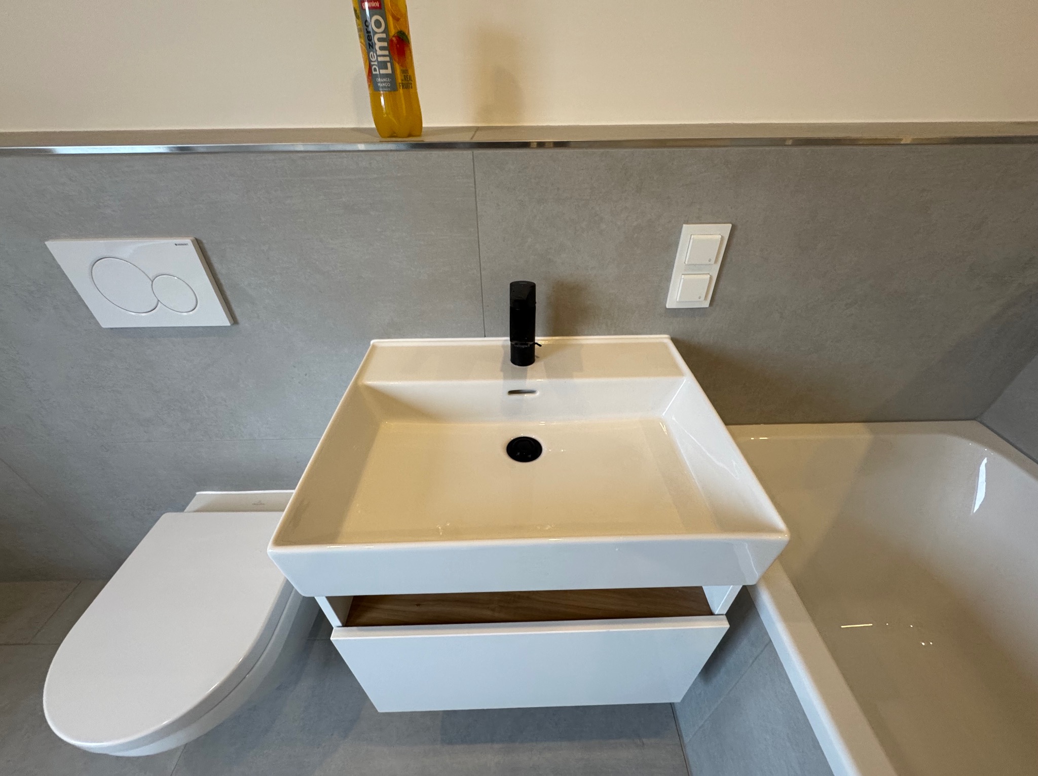 Nowoczesna łazienka z białą umywalką nablatową, toaletą i wanną. Szare płytki na ścianach, minimalistyczna armatura. Butelka z napojem na półce dodaje koloru.