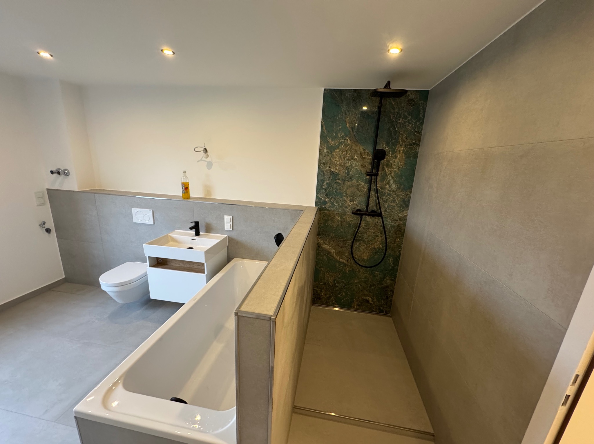 Nowoczesna łazienka z wanną, kabiną prysznicową typu walk-in z zielonym akcentem, umywalką i toaletą. Minimalistyczny design, szare płytki, oświetlenie punktowe.