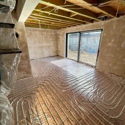 INSTALCO - Wnętrze w trakcie montażu ogrzewania podłogowego: rury ułożone na folii z siatką, surowy tynk na ścianach, widoczne drewniane belki stropu i okno przesuwne.