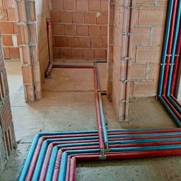 INSTALCO - Instalacja ogrzewania podłogowego w trakcie realizacji w Nowym Sączu, widoczne rury czerwone i niebieskie ułożone na podłodze w surowym stanie, ściany z cegły.