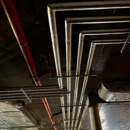 INSTALCO - Widok na instalacje pod sufitem: rury grzewcze z izolacją aluminiową układające się w kąty proste, rury z tworzywa w kolorze ceglastym oraz metalowe szyny montażowe.