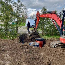 INSTALCO - Czerwona koparka Kubota umieszcza betonowy zbiornik w wykopie na tle pochmurnego nieba i białego vana, z pracownikiem w pomarańczowych rękawicach obserwującym proces.