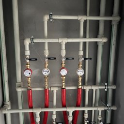 INSTALCO - Rozdzielacz instalacji wodnej z czterema wodomierzami, rurami w kolorach białym i czerwonym, zamontowany na szarej ścianie.