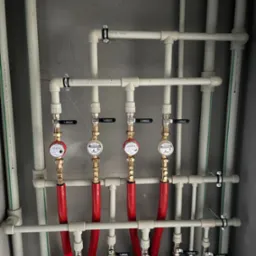 Rozdzielacz instalacji wodnej z czterema wodomierzami, rurami w kolorach białym i czerwonym, zamontowany na szarej ścianie.