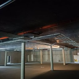 INSTALCO - Surowy stan piwnicy z widocznymi instalacjami pod sufitem, w tym rurami wentylacyjnymi i hydraulicznymi, oraz betonowymi kolumnami owiniętymi folią zabezpieczającą.