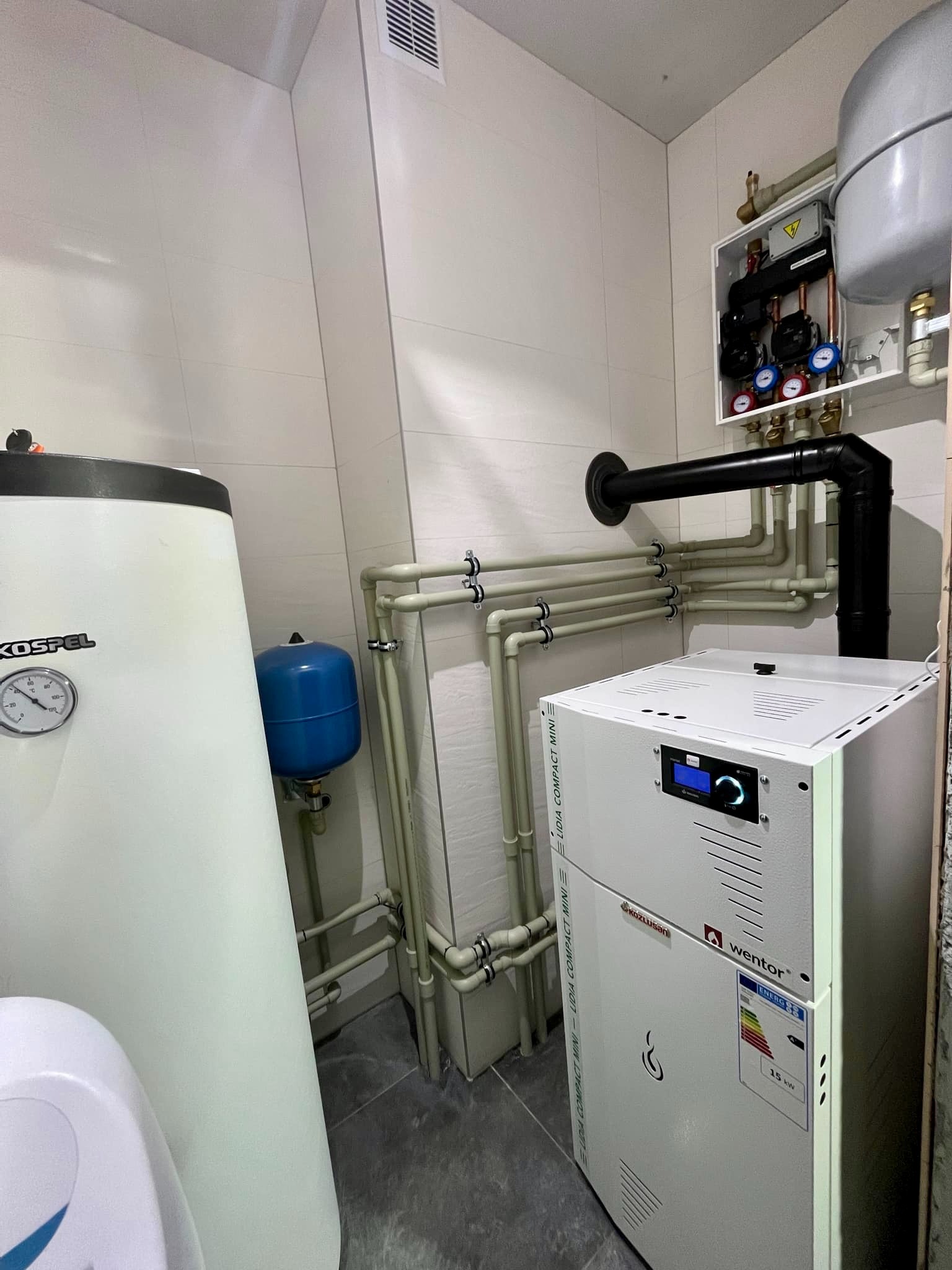 Wnętrze kotłowni z piecem CO Wentor Lidia Compact Mini, zasobnikiem Kospel, instalacją rurową i osprzętem na ścianie, w pomieszczeniu wykończonym białymi płytkami.