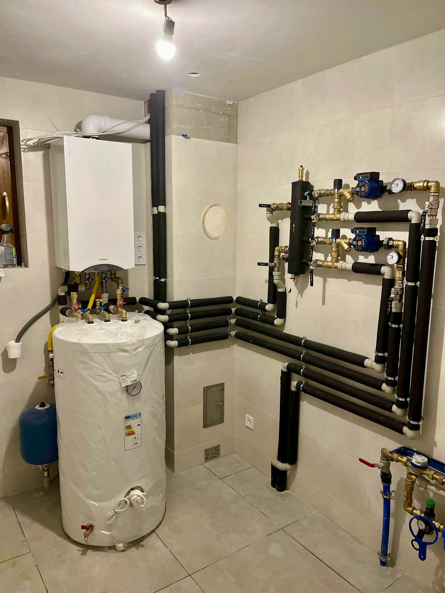 Kotłownia z białym piecem, zbiornikiem owiniętym folią i rozbudowaną instalacją rur z izolacją termiczną na beżowych płytkach.