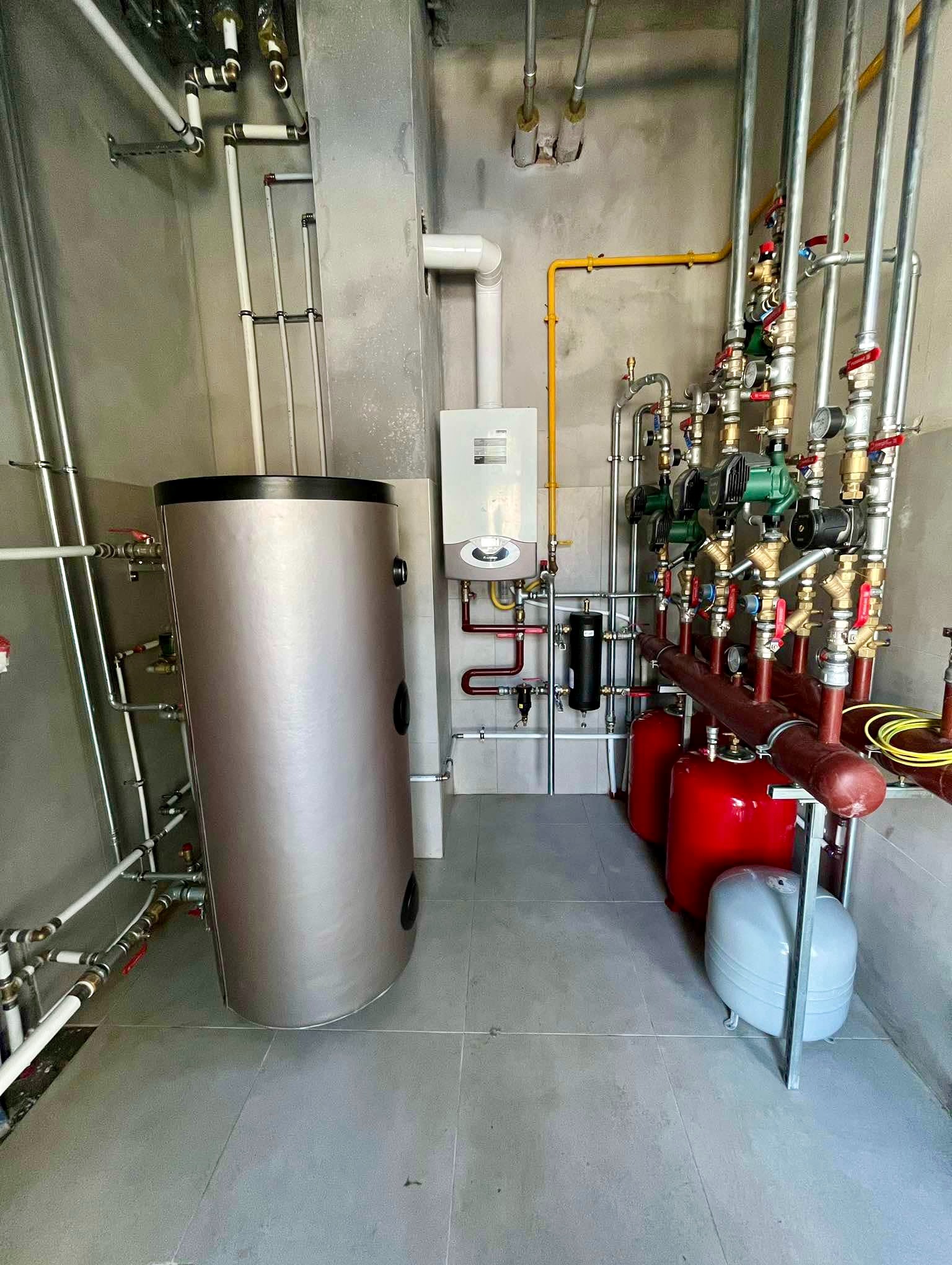 Kompletna kotłownia z szarym zbiornikiem ciepłej wody, białym piecem gazowym, czerwonymi zbiornikami wyrównawczymi i rozbudowaną instalacją rurową z licznymi zaworami i manometrami na szarych...