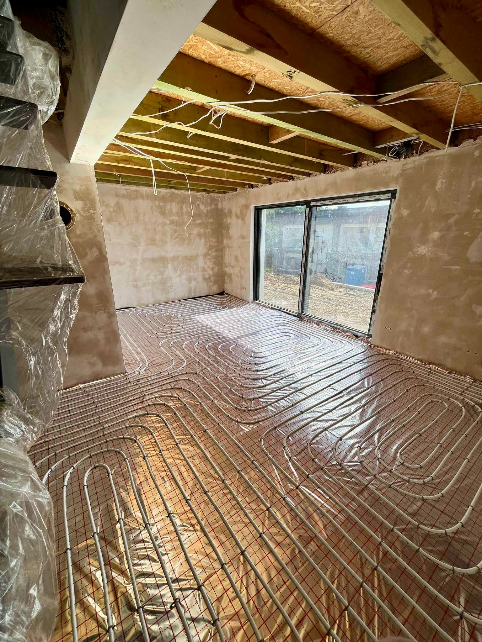 Wnętrze w trakcie montażu ogrzewania podłogowego: rury ułożone na folii z siatką, surowy tynk na ścianach, widoczne drewniane belki stropu i okno przesuwne.