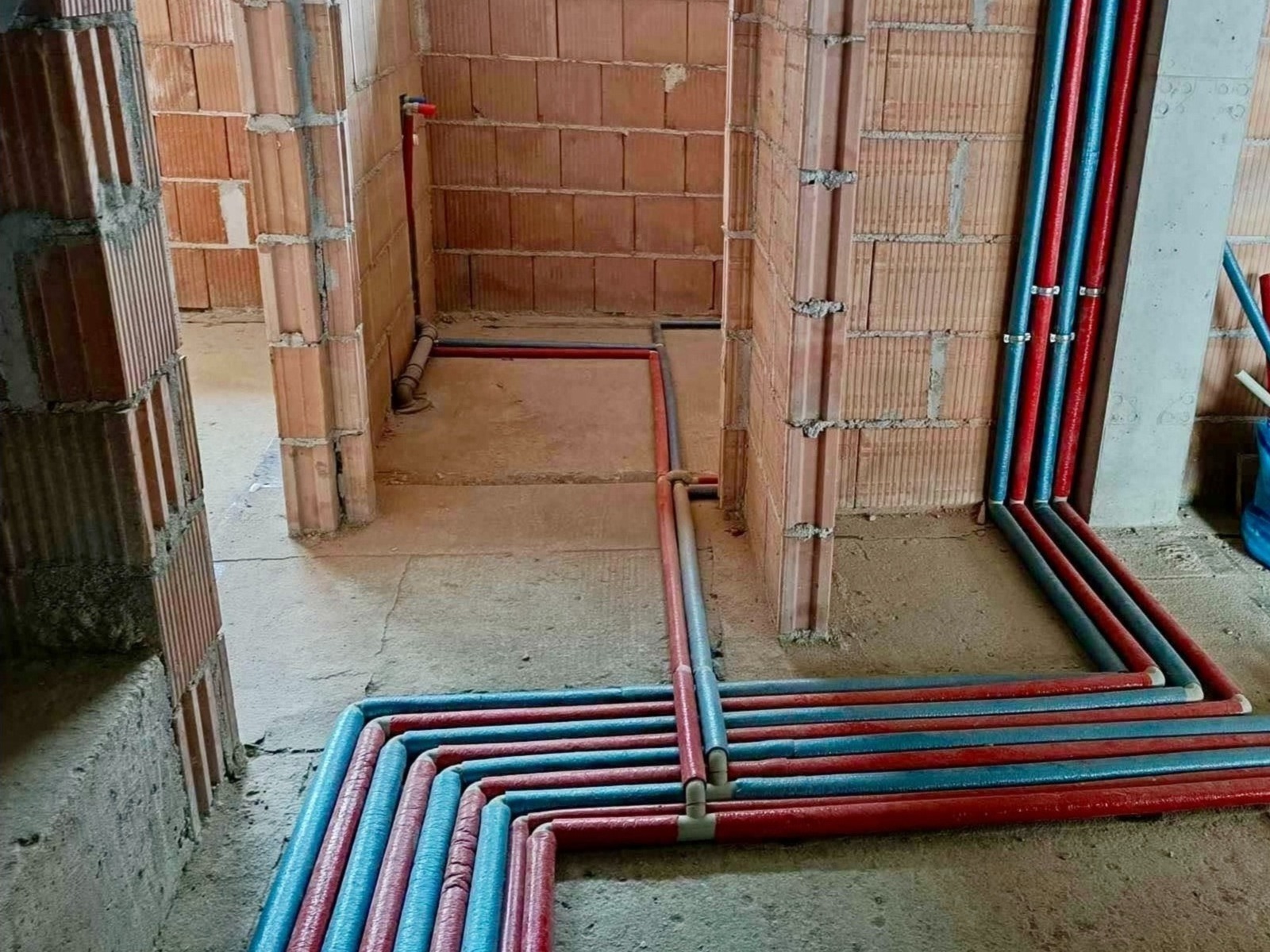 Instalacja ogrzewania podłogowego w trakcie realizacji w Nowym Sączu, widoczne rury czerwone i niebieskie ułożone na podłodze w surowym stanie, ściany z cegły.