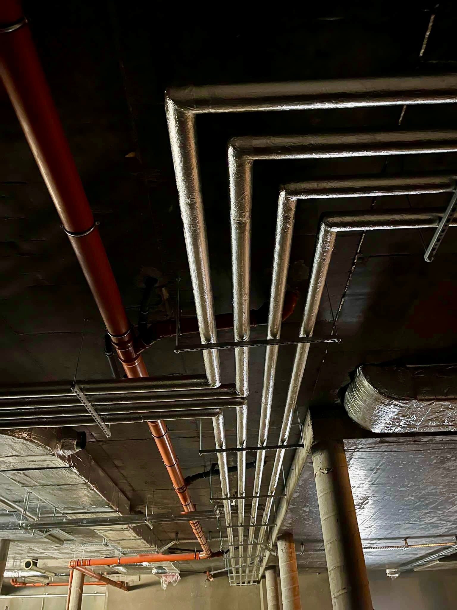 Widok na instalacje pod sufitem: rury grzewcze z izolacją aluminiową układające się w kąty proste, rury z tworzywa w kolorze ceglastym oraz metalowe szyny montażowe.