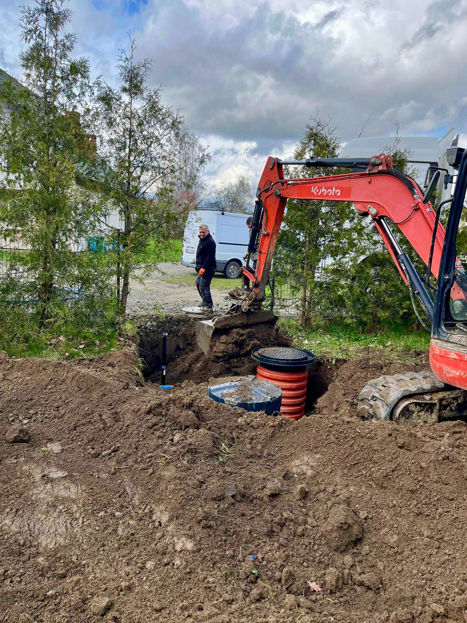 Czerwona koparka Kubota umieszcza betonowy zbiornik w wykopie na tle pochmurnego nieba i białego vana, z pracownikiem w pomarańczowych rękawicach obserwującym proces.