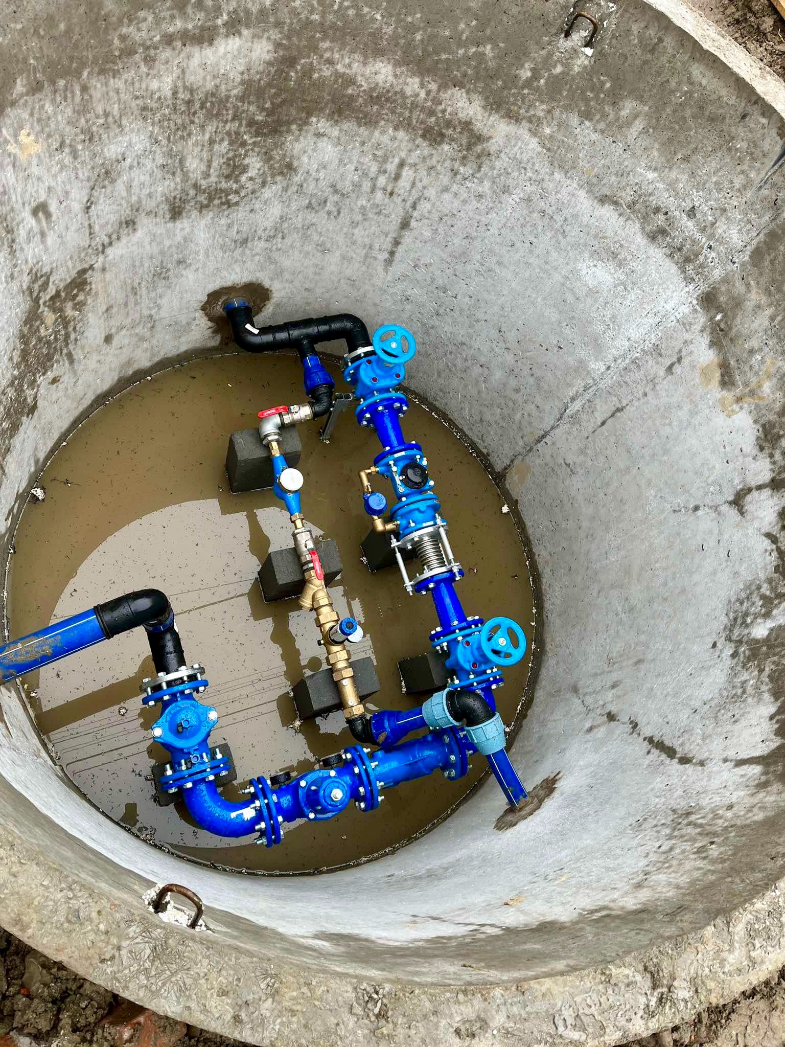 Wnętrze betonowego kręgu studni z zainstalowanymi niebieskimi rurami i zaworami, częściowo zanurzonymi w wodzie, widoczne czarne rury i metalowe uchwyty w ścianie studni.