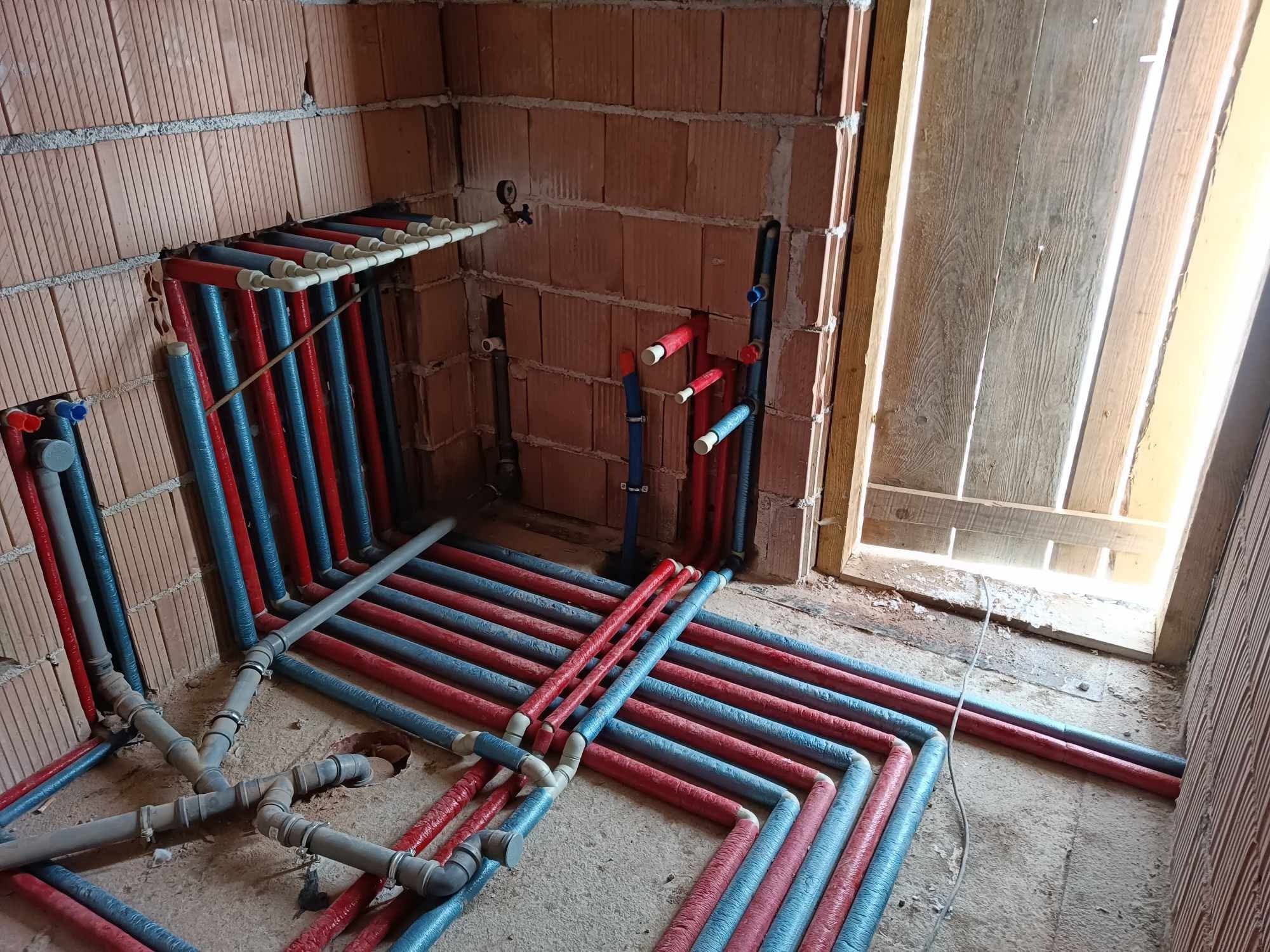 Wnętrze w trakcie prac hydraulicznych: liczne rury w izolacji czerwonej i niebieskiej, prowadzone po podłodze i ścianach z cegły. Widoczne złącza i kolanka, fragment drewnianej ościeżnicy.