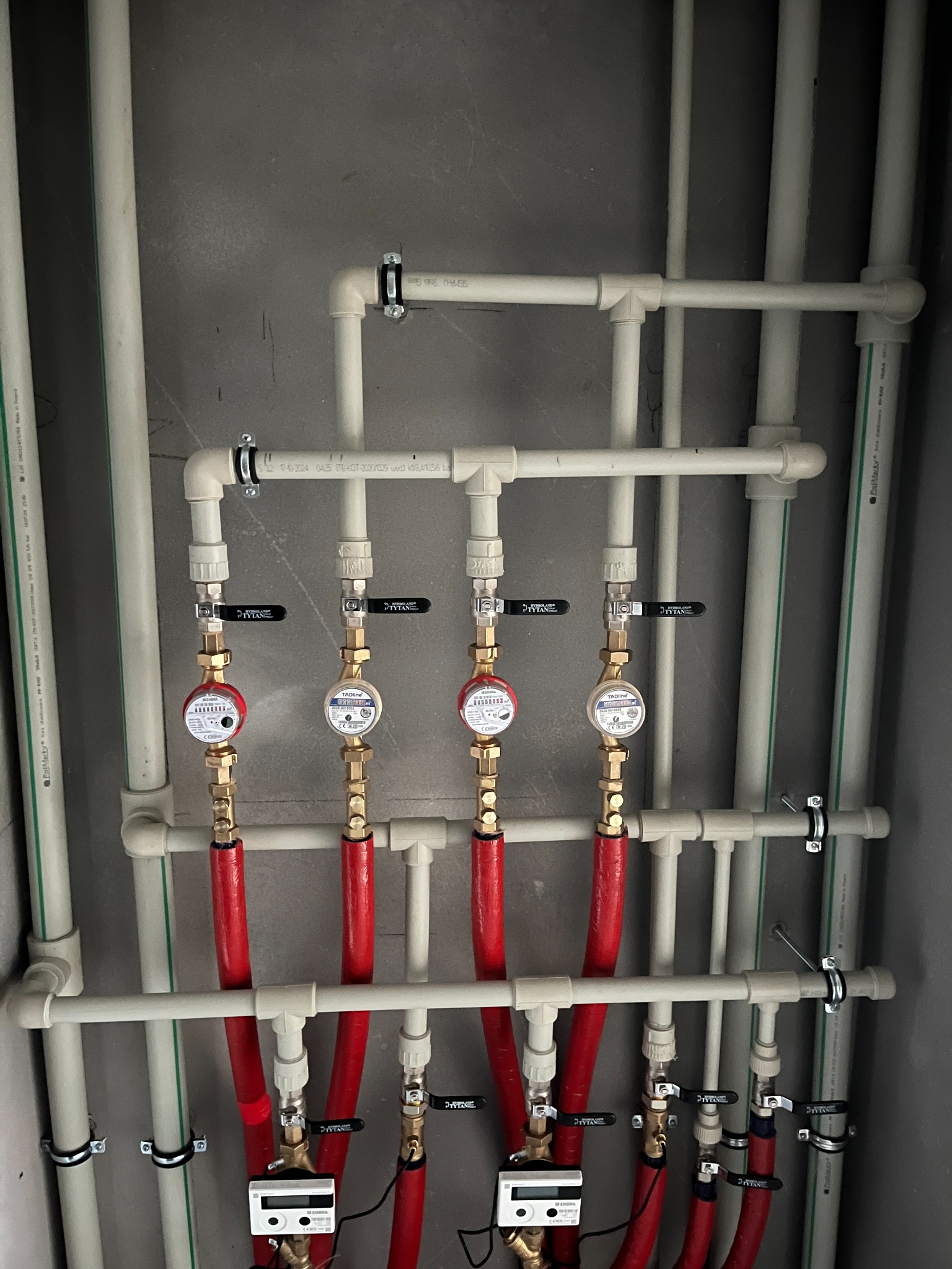 Rozdzielacz instalacji wodnej z czterema wodomierzami, rurami w kolorach białym i czerwonym, zamontowany na szarej ścianie.