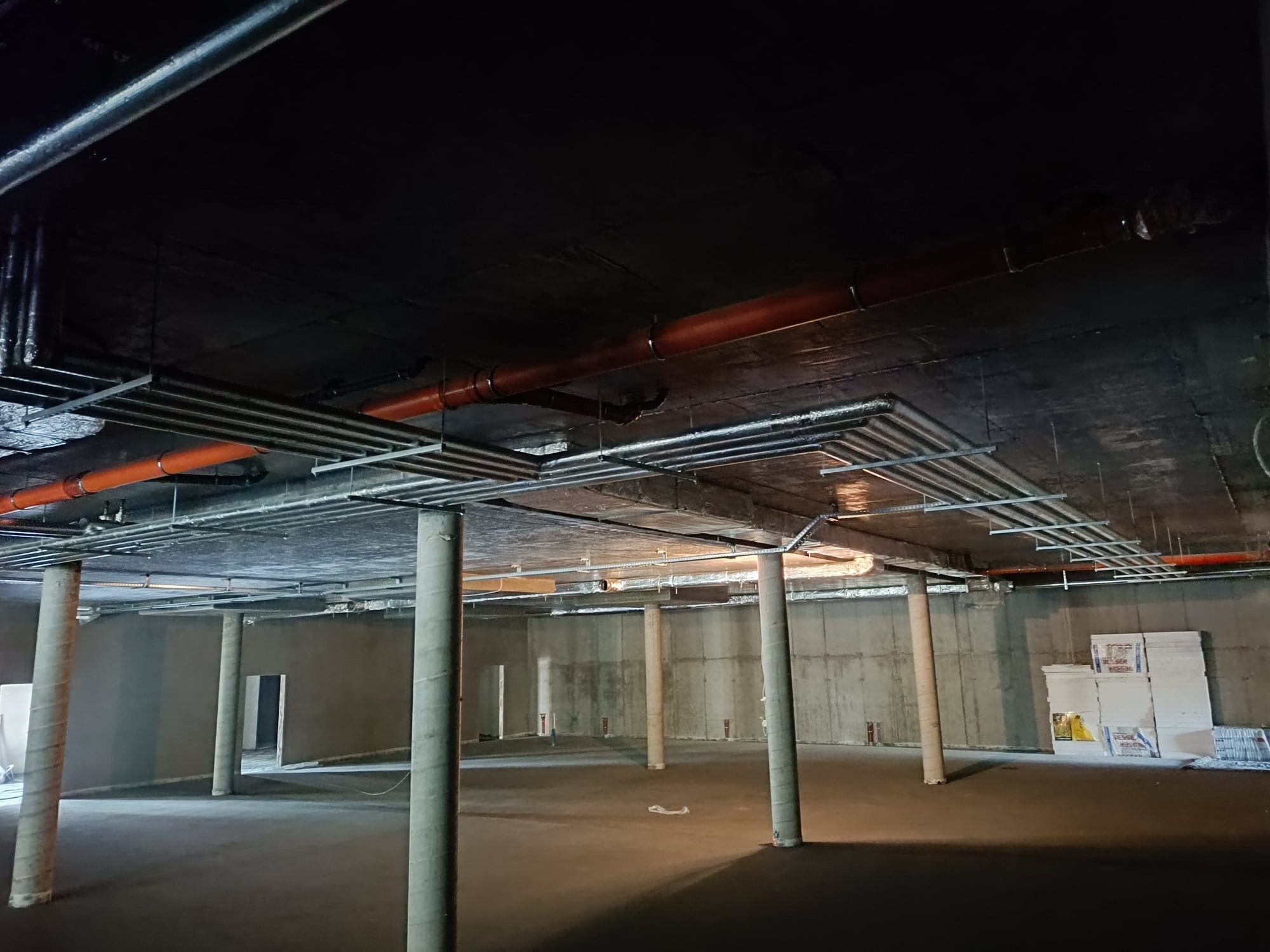Surowy stan piwnicy z widocznymi instalacjami pod sufitem, w tym rurami wentylacyjnymi i hydraulicznymi, oraz betonowymi kolumnami owiniętymi folią zabezpieczającą.