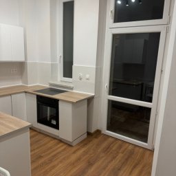 VS GROUPE OLEKSANDR PETRENKO - Nowoczesna kuchnia z białymi szafkami, drewnianymi blatami i podłogą z paneli imitujących drewno, widoczny fragment okna i drzwi balkonowych.