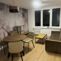 Jasne wnętrze salonu z okrągłym stołem, krzesłami z tapicerką w odcieniach beżu, brązową sofą i lampą wiszącą z drewnianym akcentem, na podłodze panele podłogowe o jasnym odcieniu drewna, ściana...