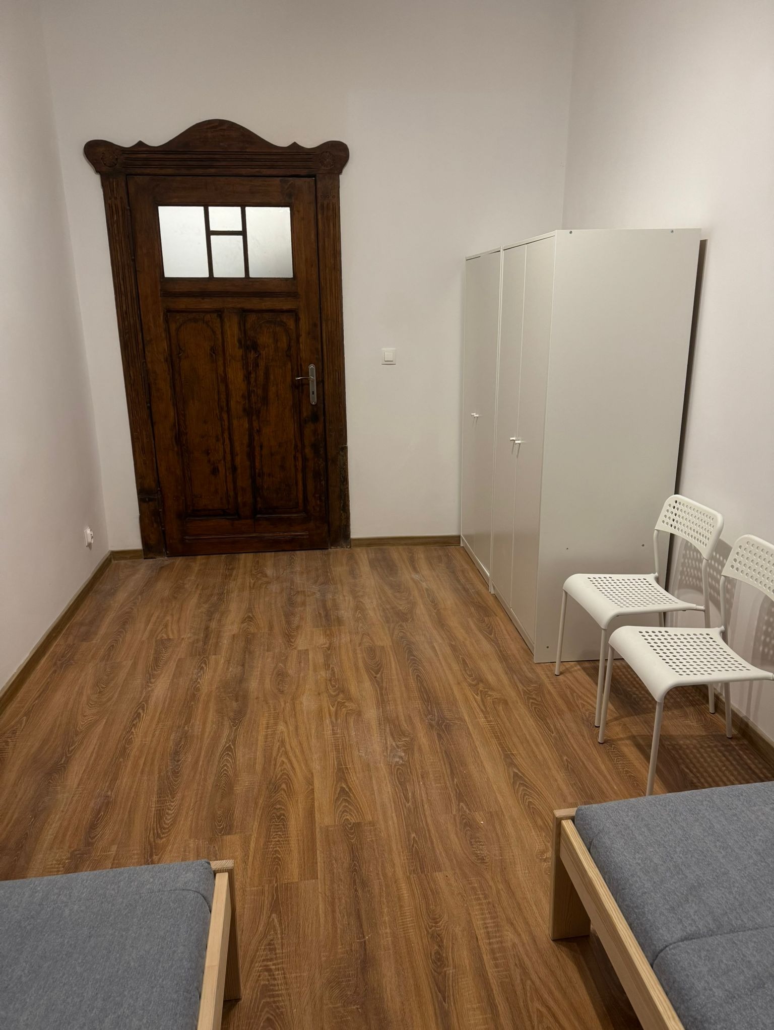 Wnętrze pokoju z drewnianą podłogą imitującą panele, ozdobnymi drzwiami z szybkami, białymi szafami i krzesłami, oraz fragmentem szarej sofy.