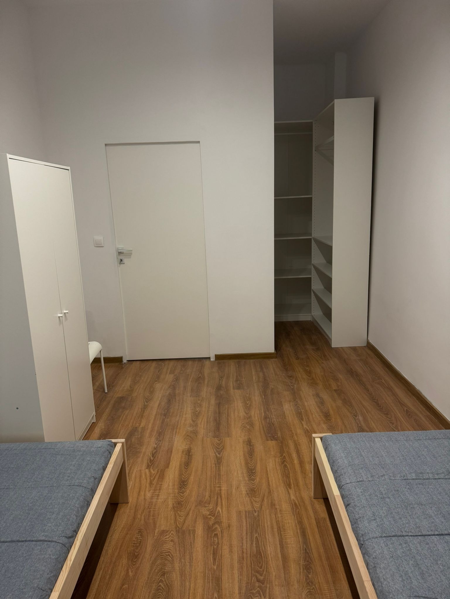 Pusty pokój z drewnianą podłogą, białymi ścianami, szafą wnękową z półkami, białym regałem i dwoma łóżkami z szarymi materacami.