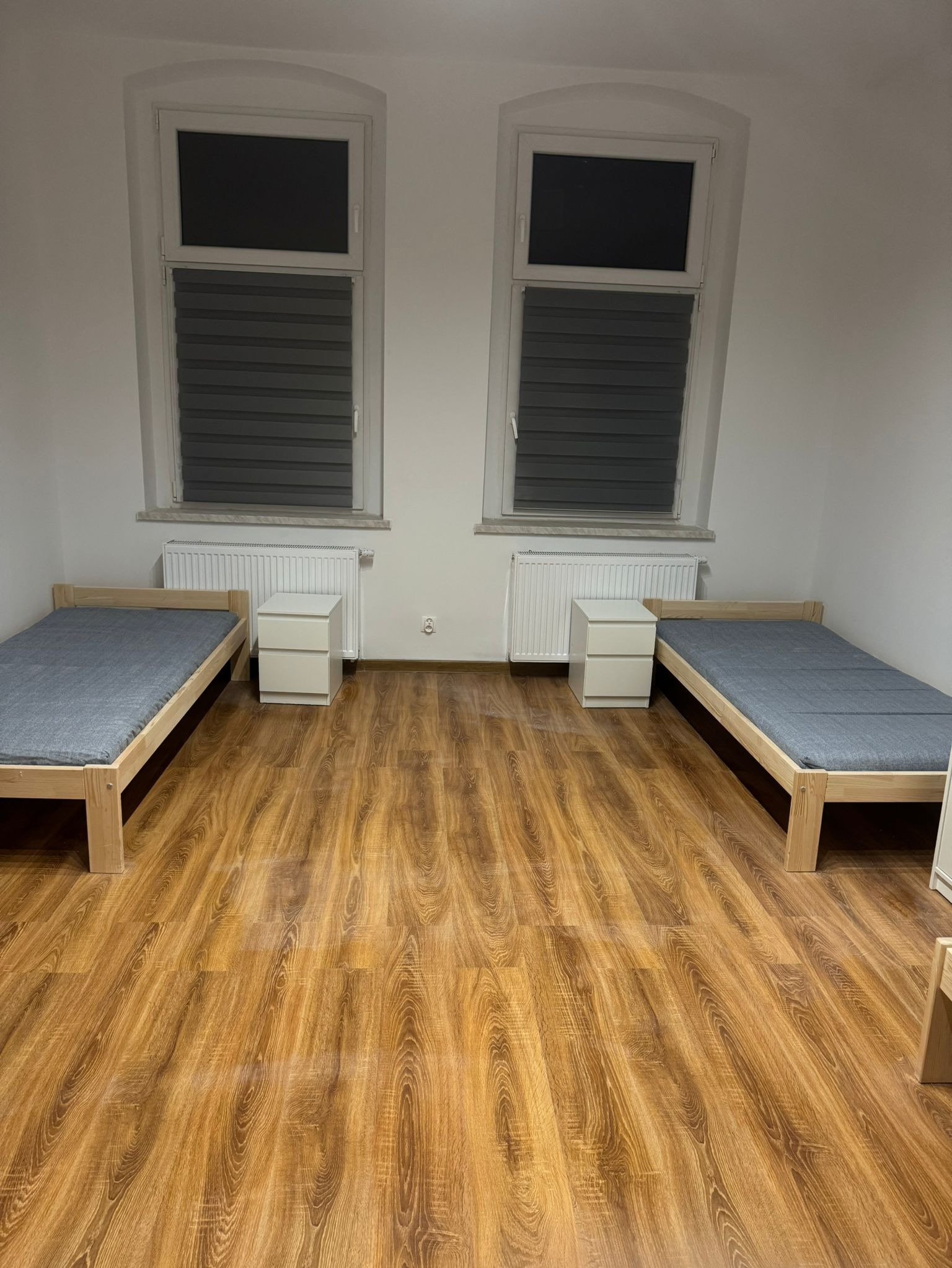 Pokój z dwoma łóżkami z jasnego drewna, szarymi materacami, białymi szafkami nocnymi i panelami podłogowymi o wzorze drewna, oświetlony przez dwa okna z żaluzjami.