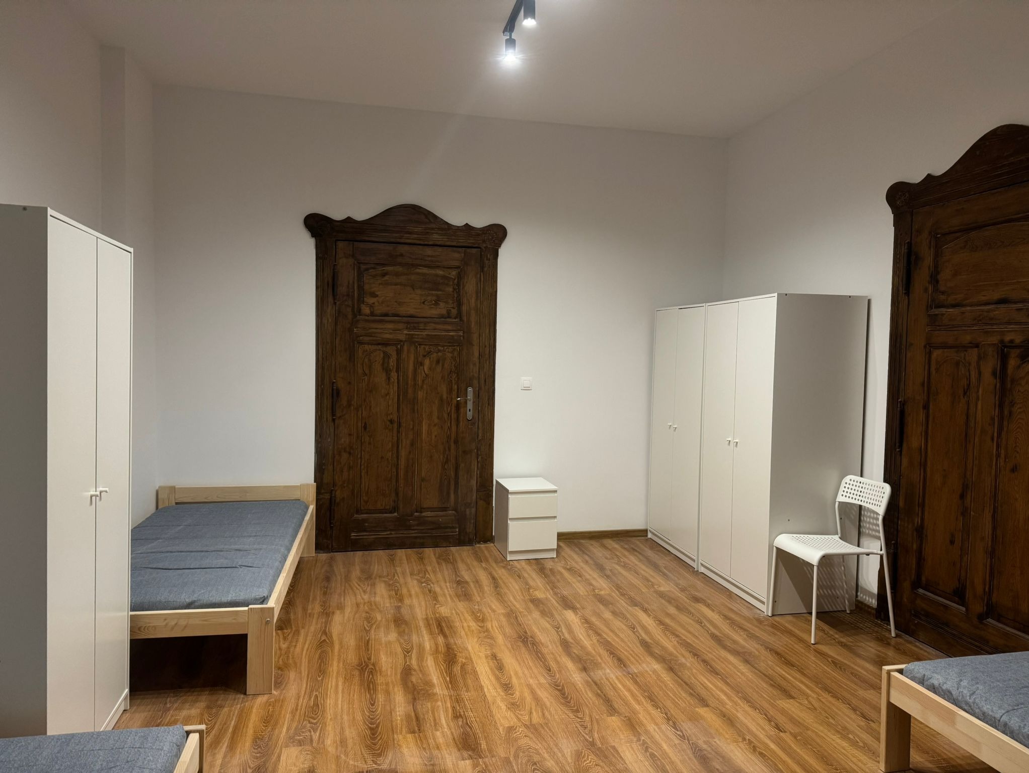 Pokój po remoncie z panelami podłogowymi, białymi szafami, drewnianymi drzwiami i łóżkami z szarymi materacami.