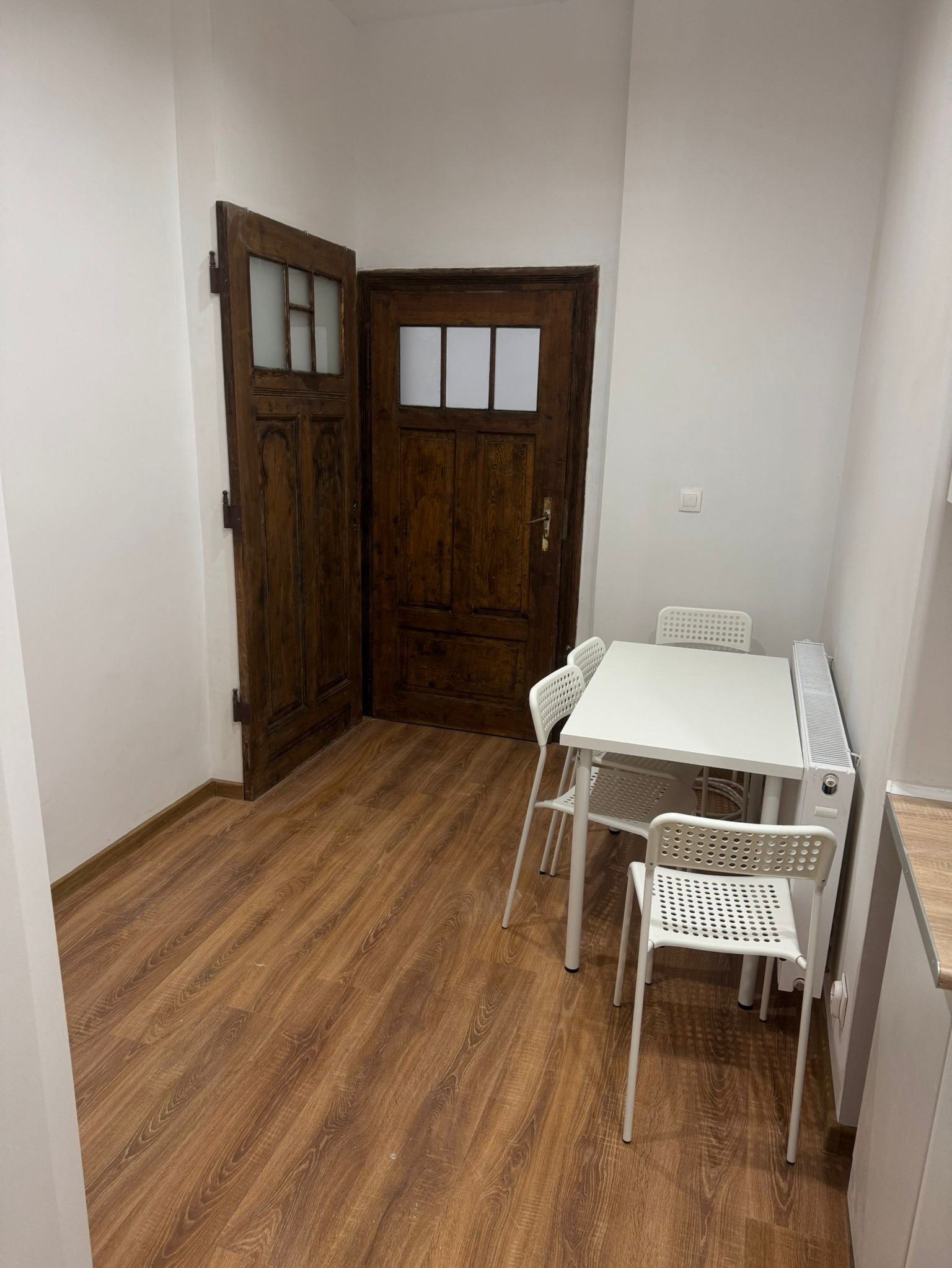 Wnętrze pokoju po remoncie: parkiet laminowany, odnowione drewniane drzwi z przeszkleniami, biały stół z krzesłami i grzejnik.